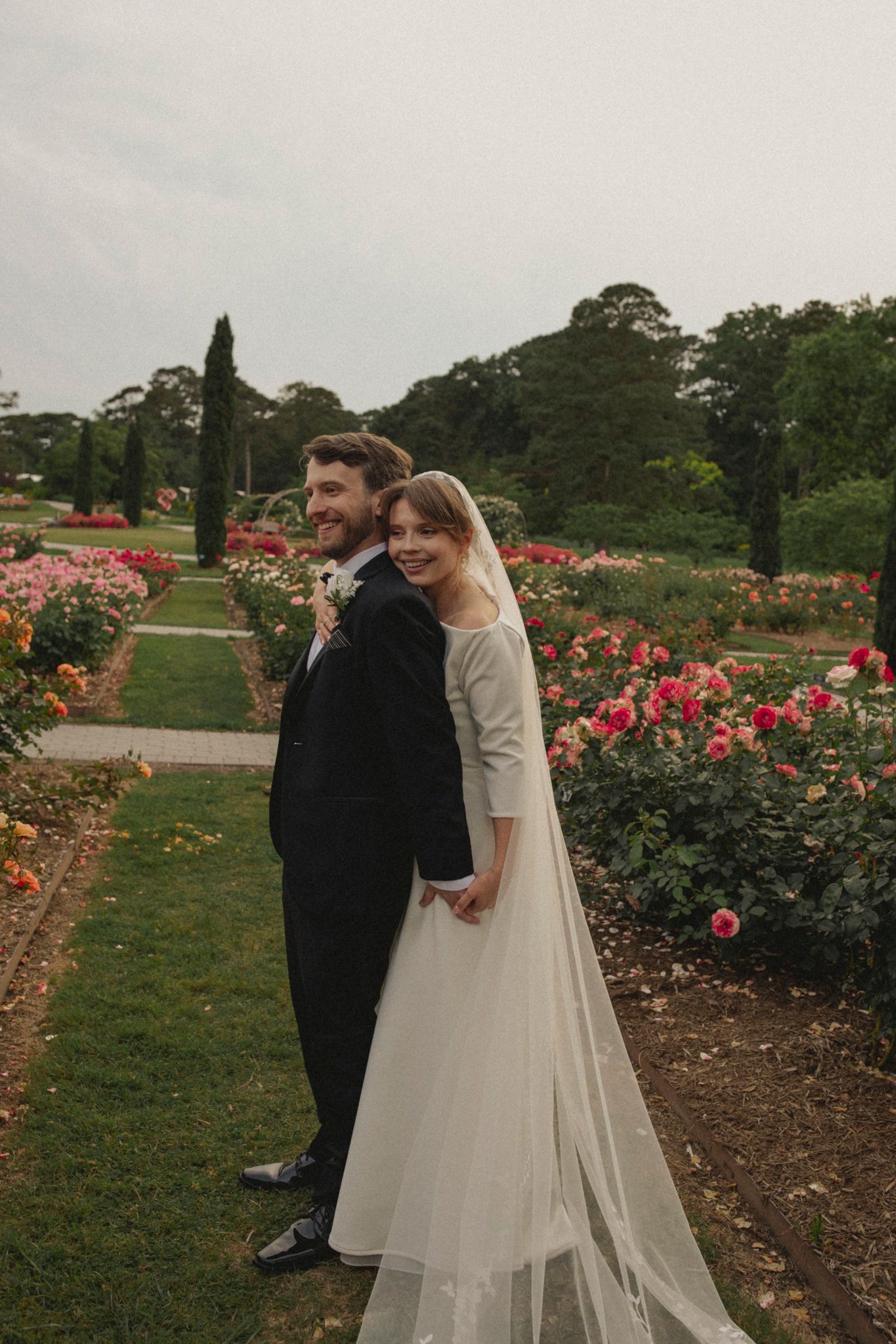 A couple in wedding attire embraces in a beautiful rose garden with blooming pink flowers and green trees in background.