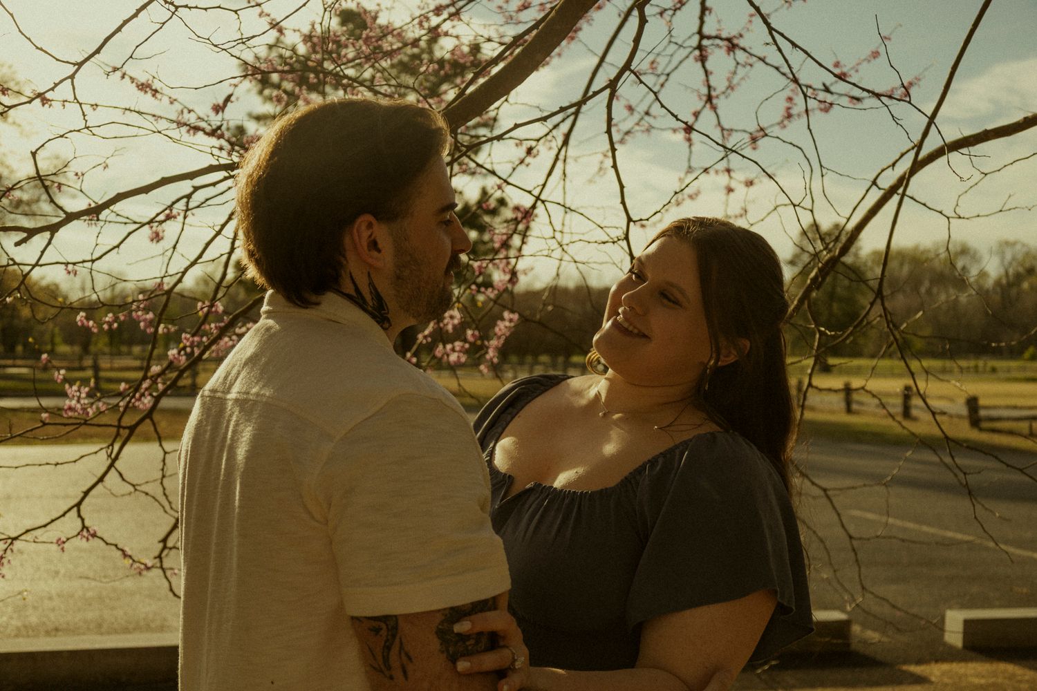 Romantic couple gazing at each other outdoors near blooming cherry blossom tree in warm golden sunlight.