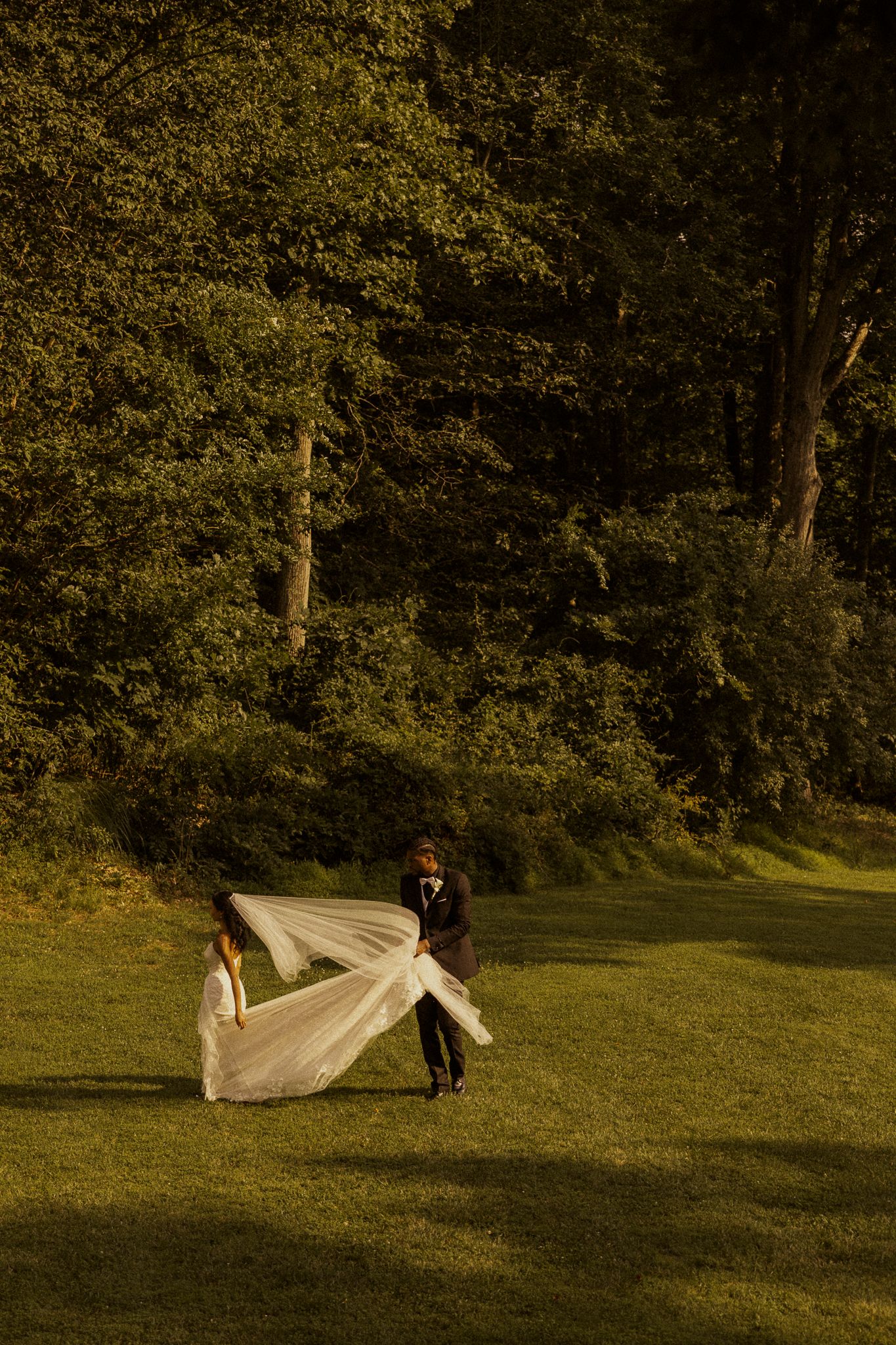 Bride and groom walking on lush green lawn surrounded by trees during golden hour wedding photography.