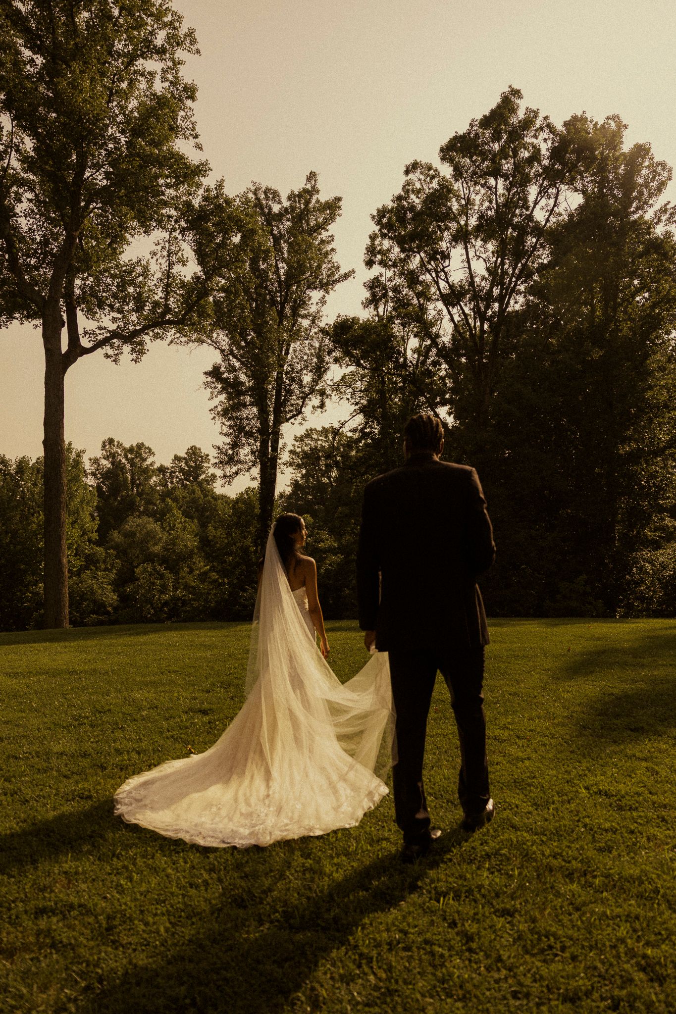 Bride and groom holding hands walking through lush green park at golden hour, long cathedral veil trailing behind.