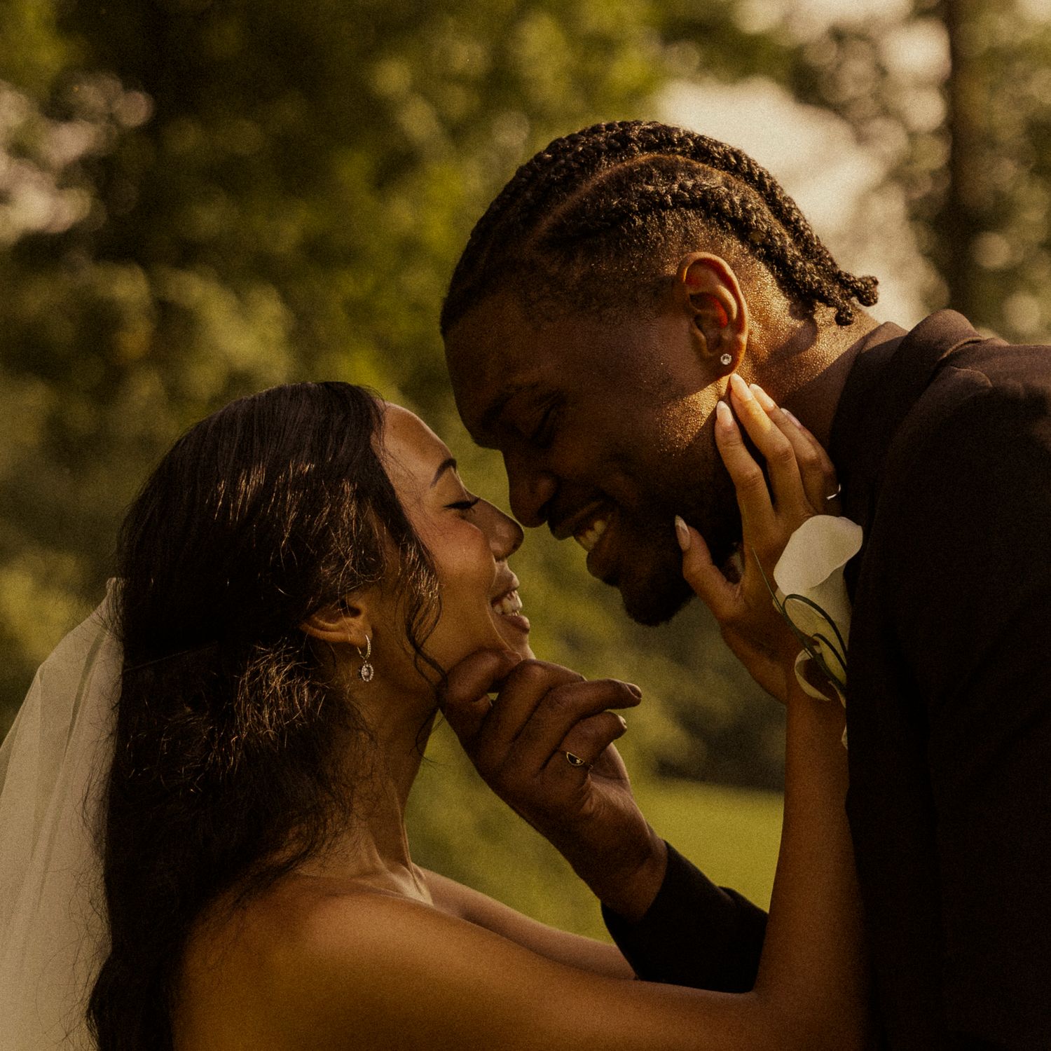 A Black bride and groom share an intimate moment, touching faces and smiling at each other outdoors in golden light.