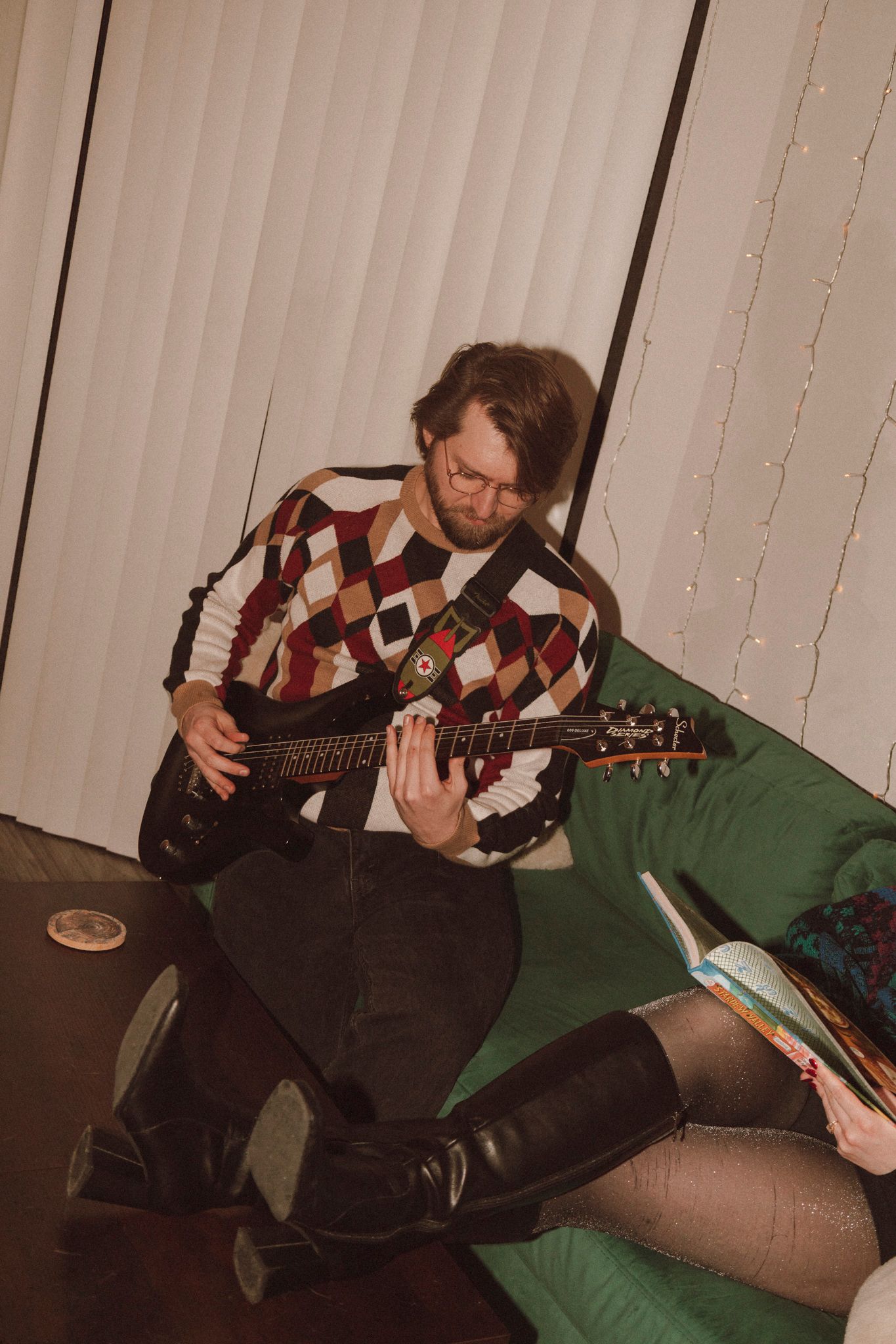 A sequence of someone playing acoustic guitar while sitting on a green couch with vertical blinds in the background.