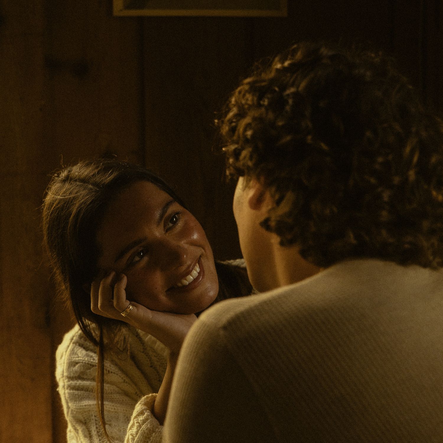 Two people share an intimate moment in warm, dim lighting, one smiling warmly at the other in a cozy wooden setting.