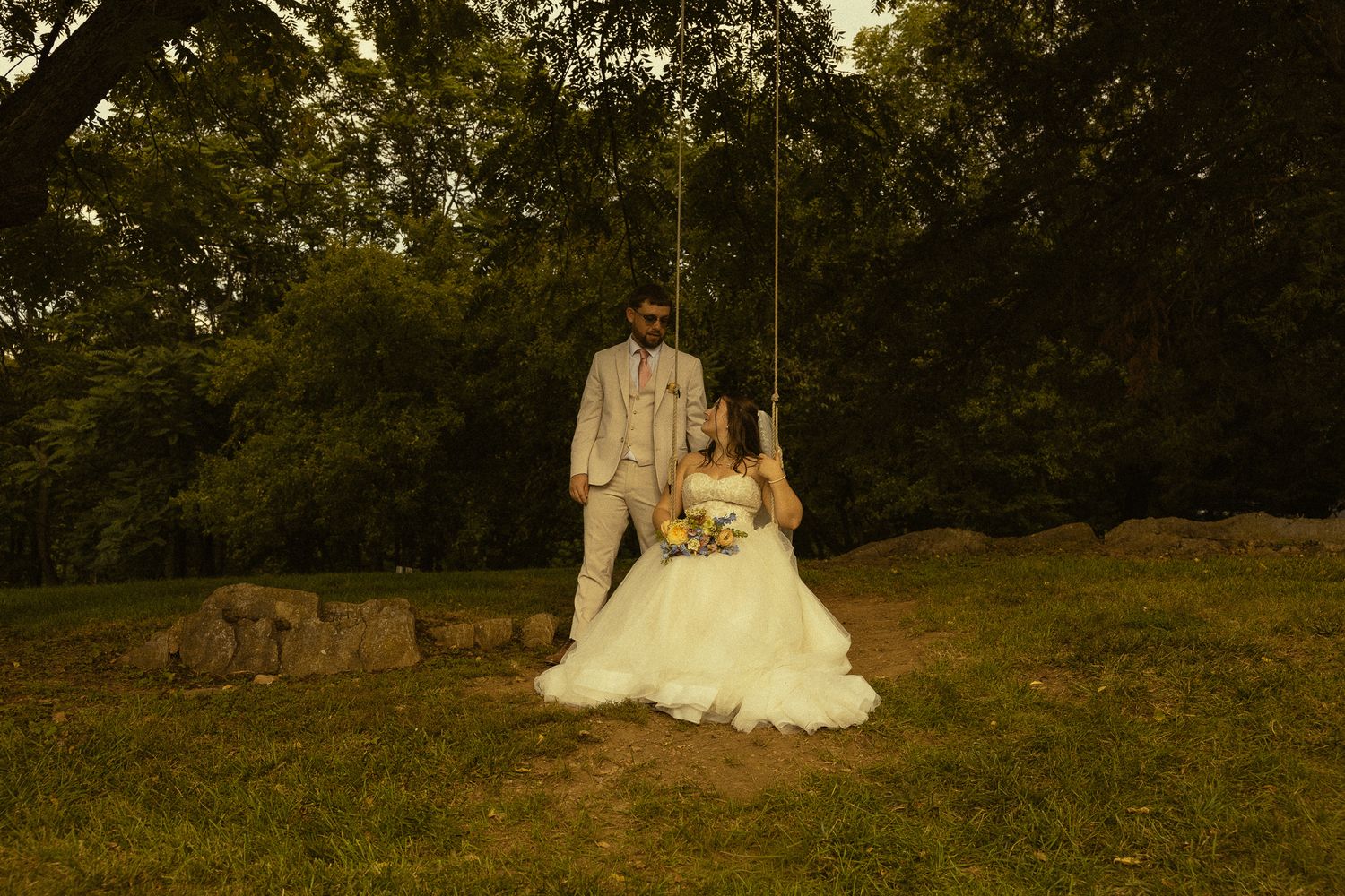 Bride sitting on a wooden swing holding a bouquet while groom stands behind her in a lush green outdoor wedding setting.