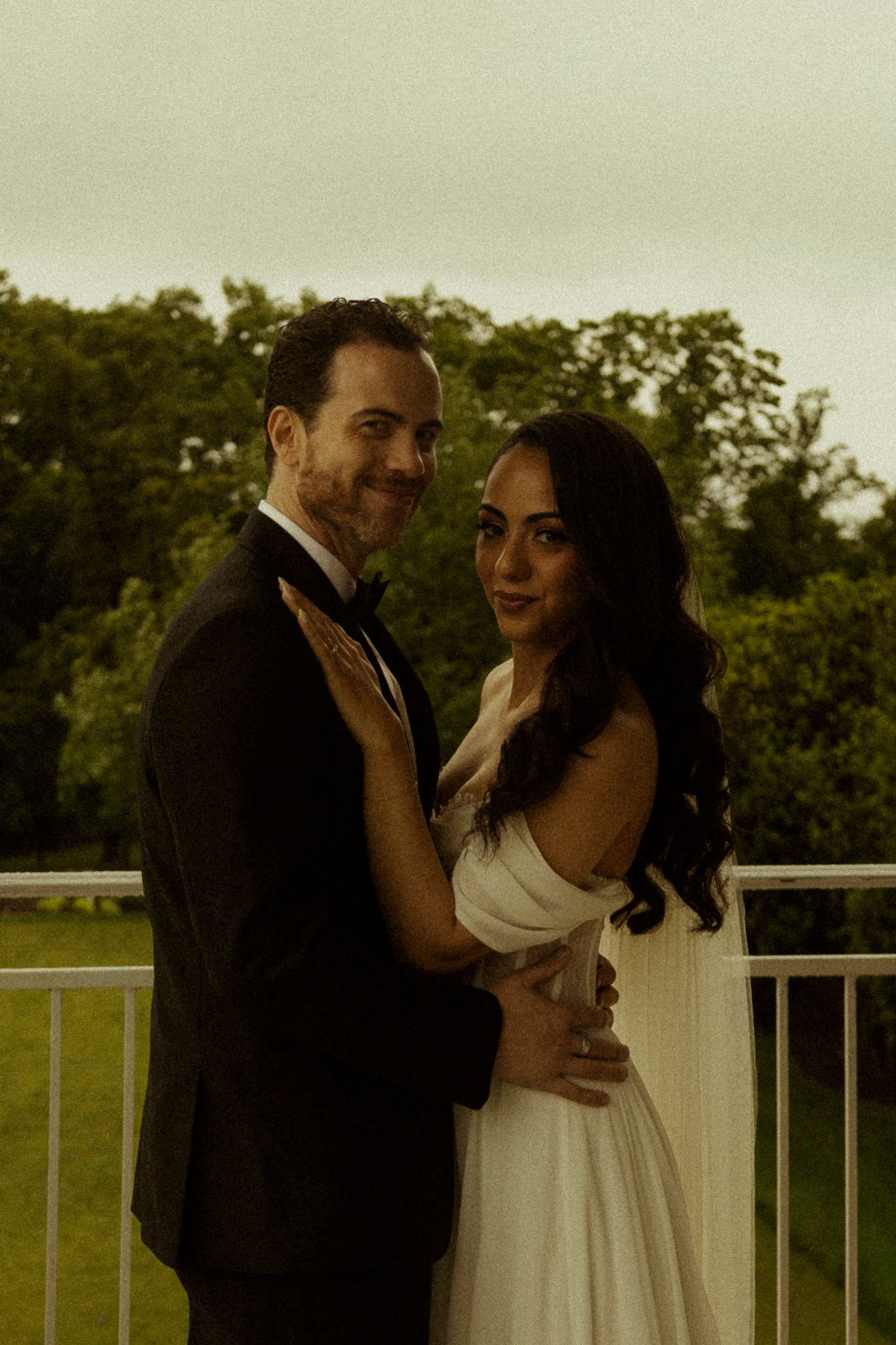 A couple in wedding attire embracing on a balcony, groom in tuxedo and bride in white gown, with lush greenery behind.