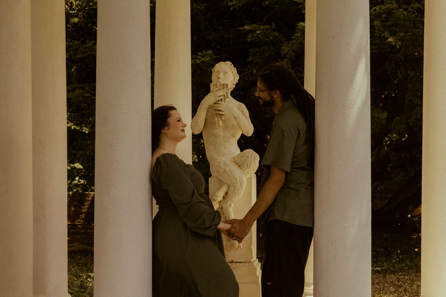 Couple holding hands between white columns with a classical marble statue in a romantic outdoor garden setting.