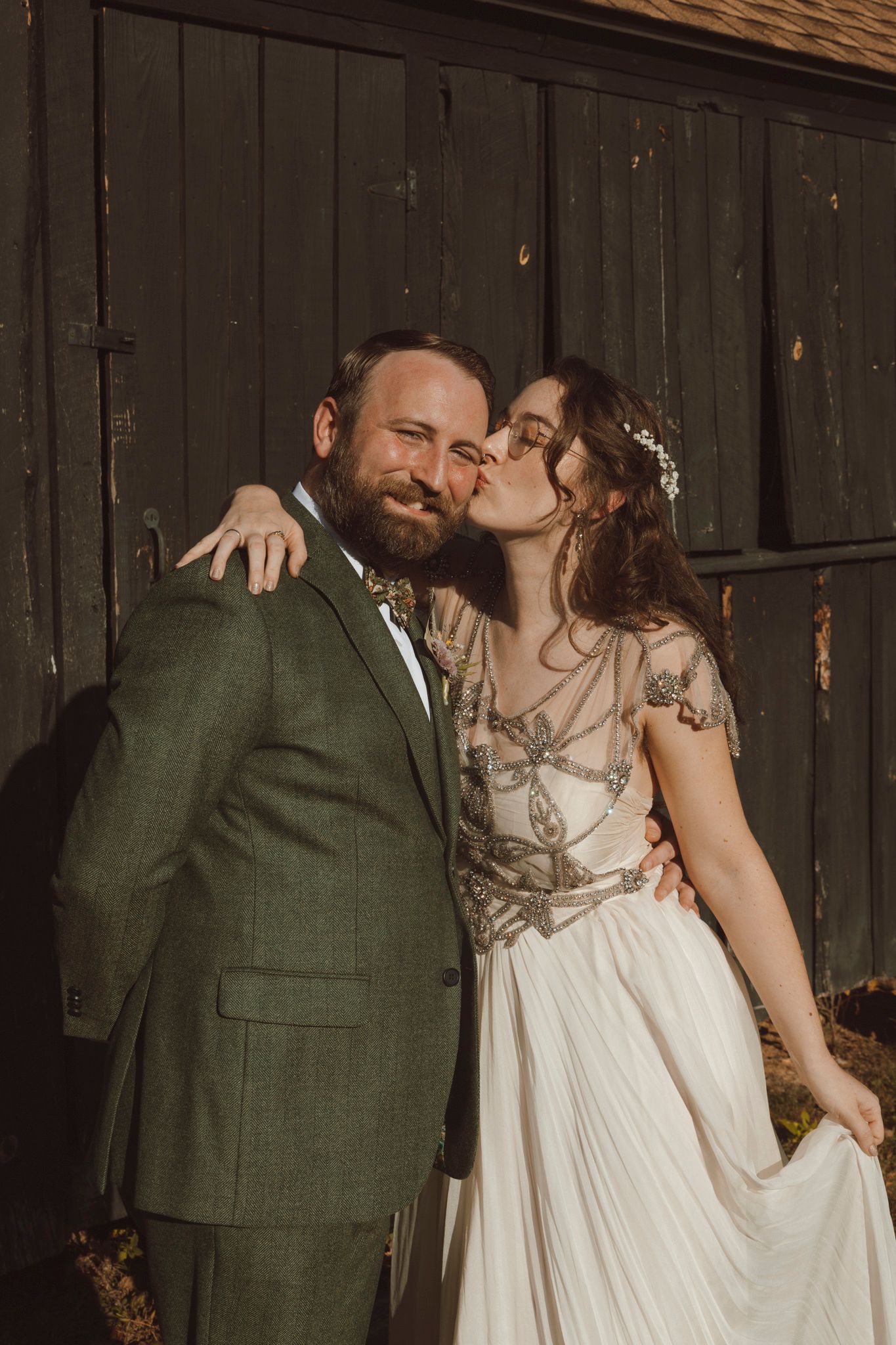 A romantic wedding couple embraces and shares a kiss in front of a rustic wooden barn wall.