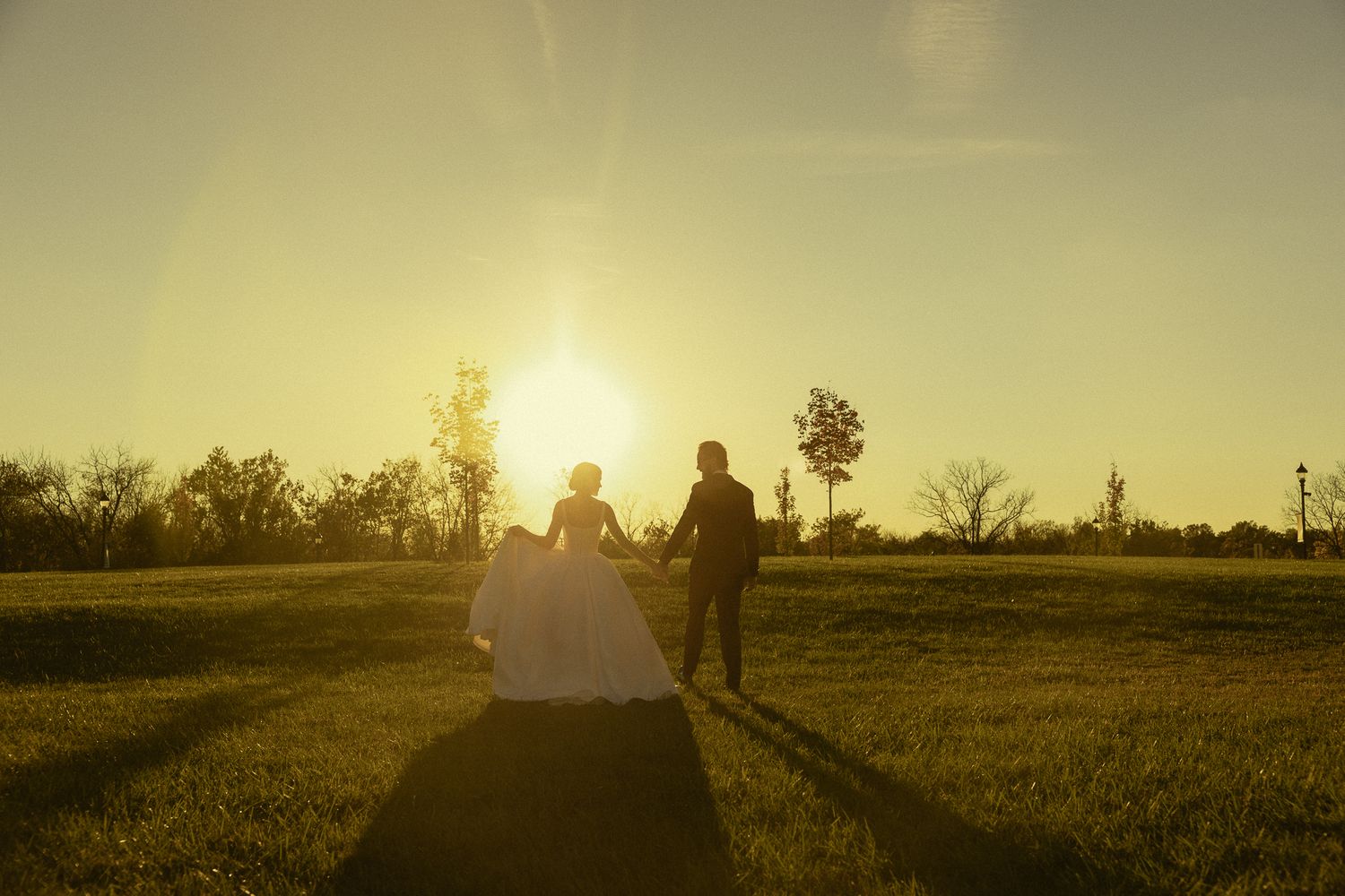 Bride in a white gown and groom in a dark suit stand on a grassy field, silhouetted by a glowing golden sunset.