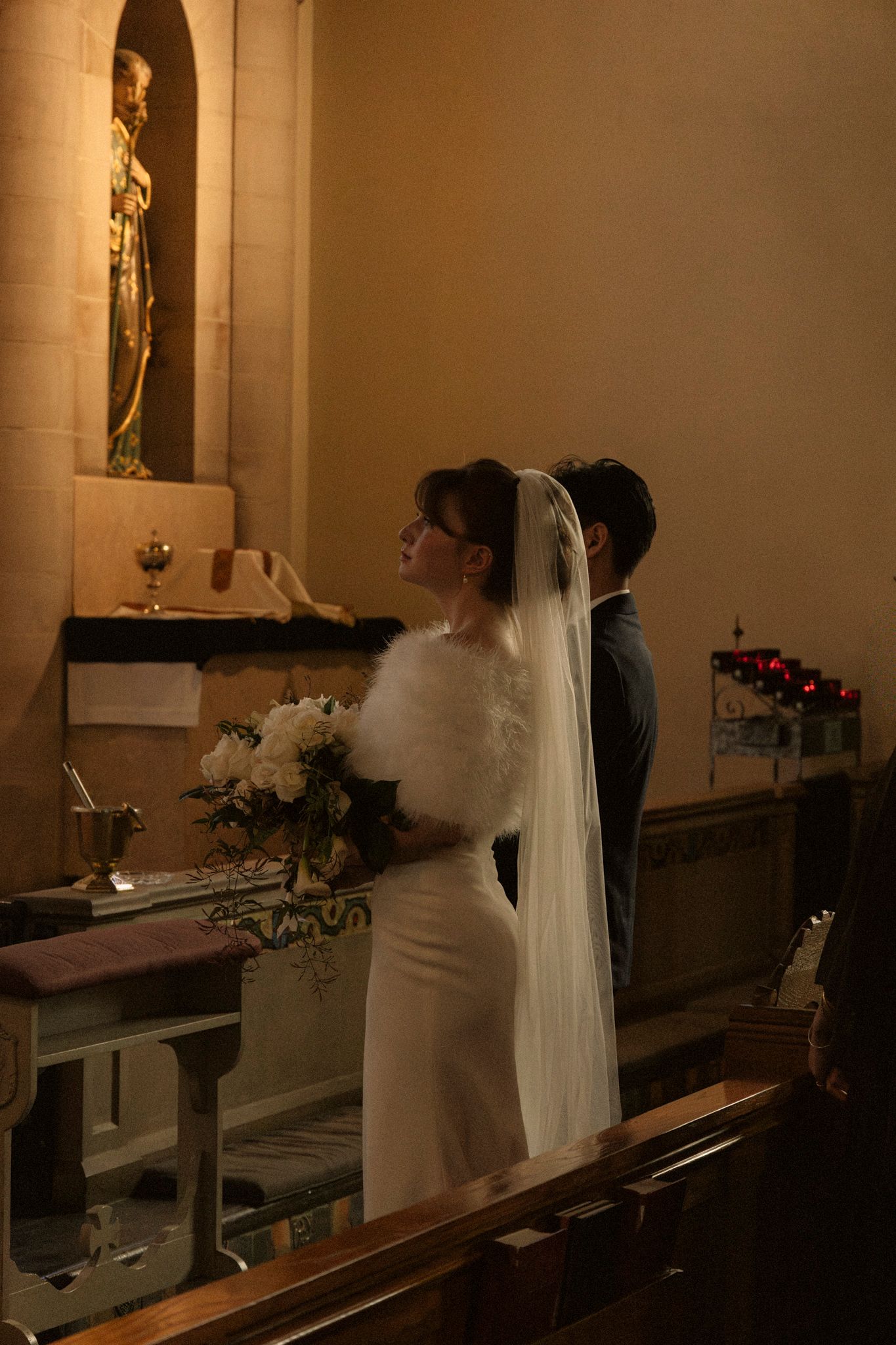 Bride in white dress and veil stands at church altar holding bouquet during traditional wedding ceremony.