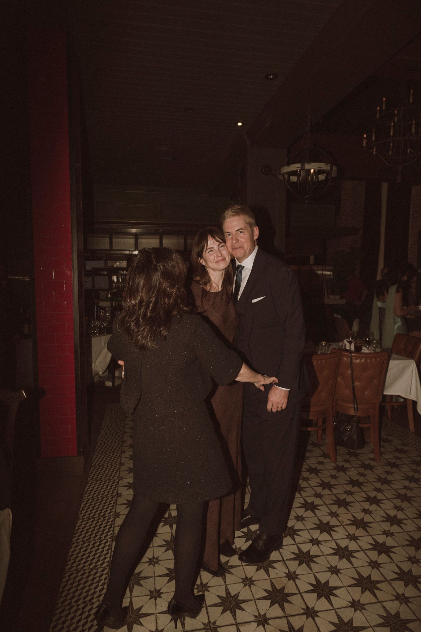 Three guests share a warm embrace while dancing on the patterned floor at a wedding reception.