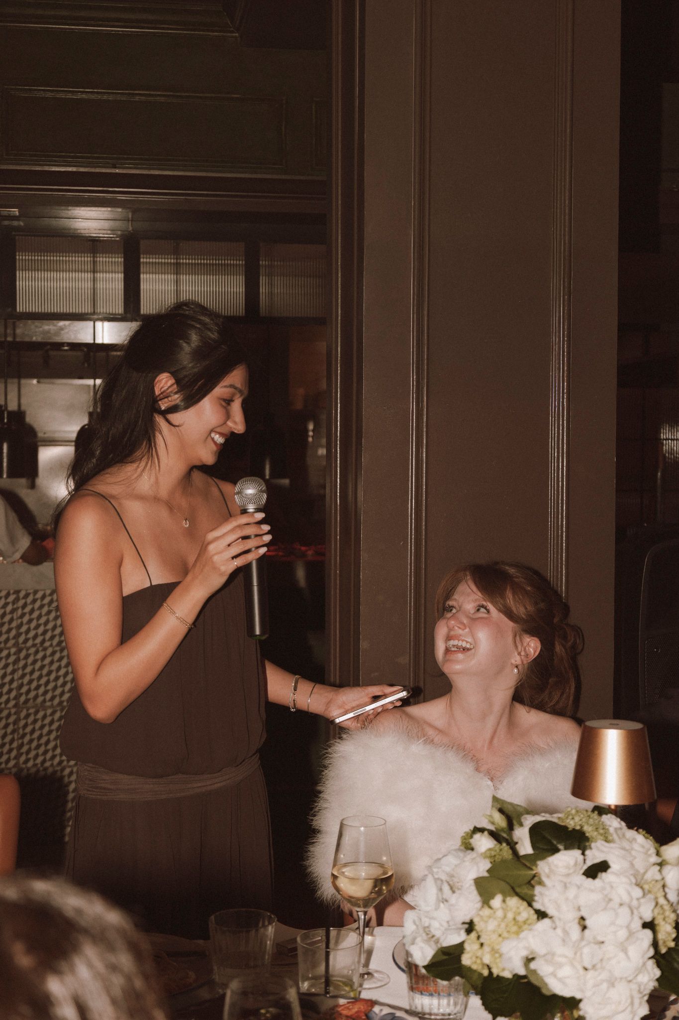 Bridesmaid giving speech with microphone while bride in white feather wrap laughs at wedding reception.