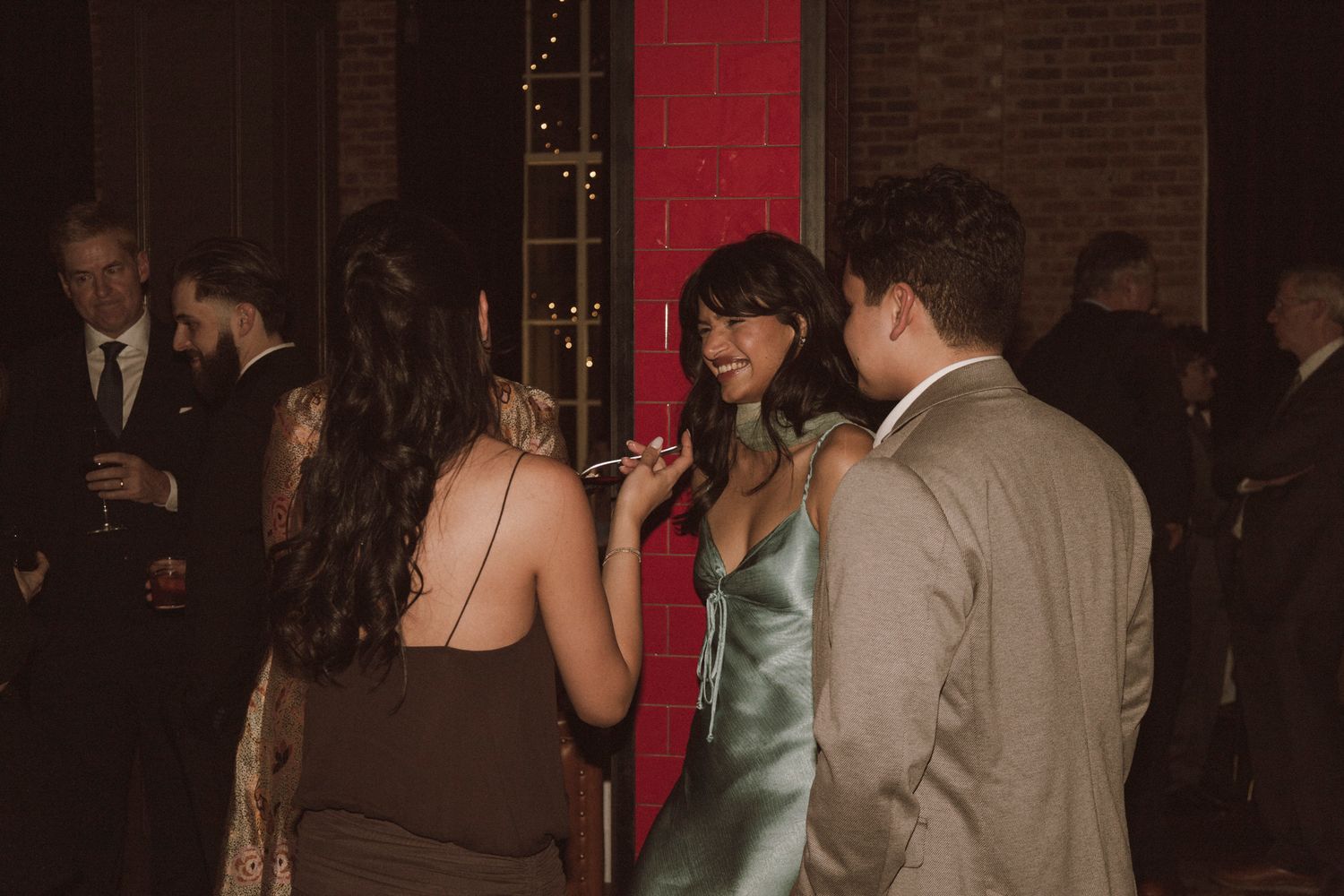 Wedding reception guests mingling in dimly lit venue with red brick wall backdrop.