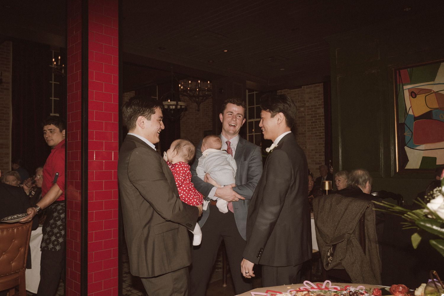 Three men in suits at a formal indoor event, one holding a baby in red, with dim restaurant lighting.