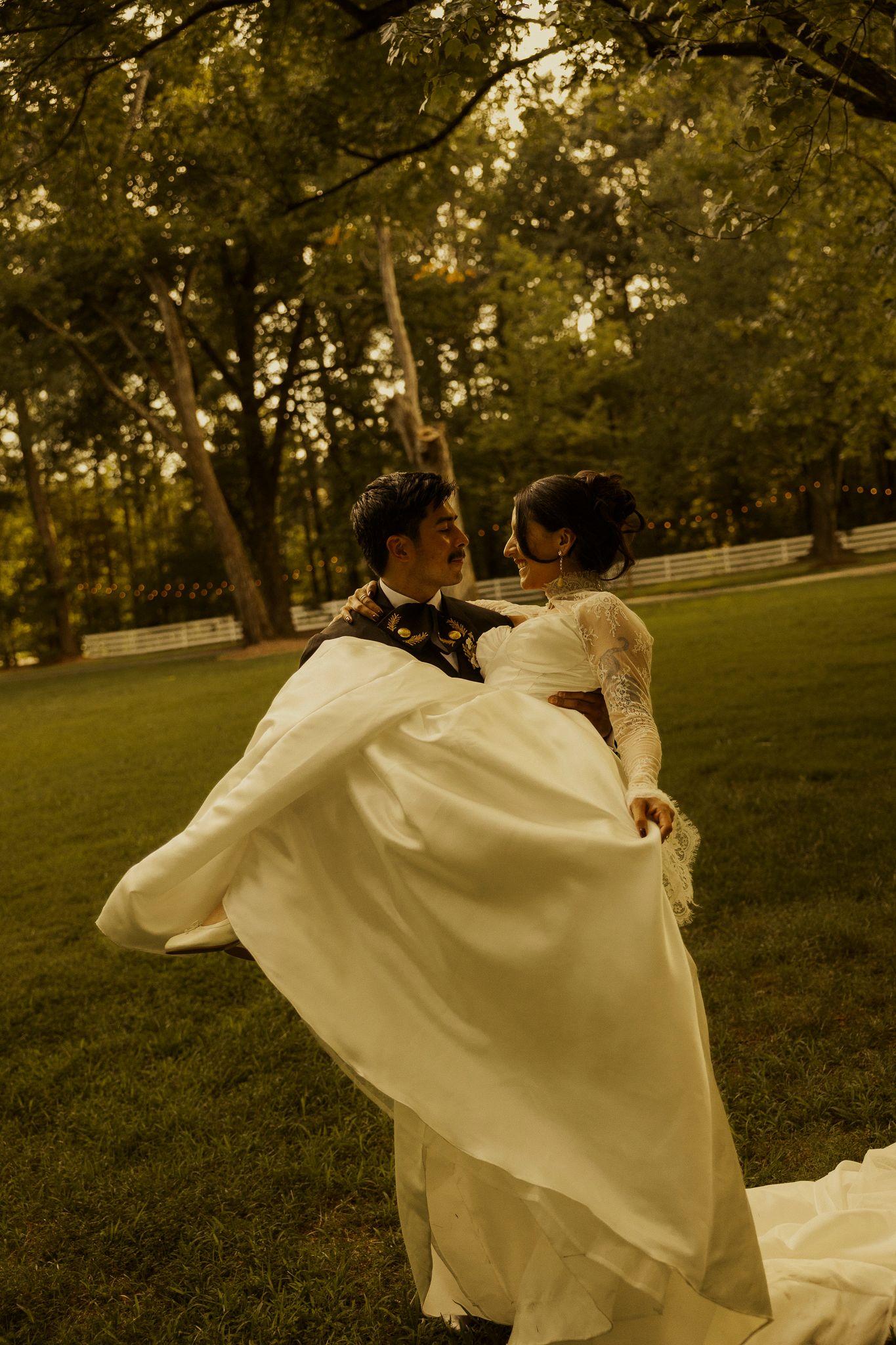 Groom carries bride in white lace gown outdoors on green lawn surrounded by trees in warm golden light.