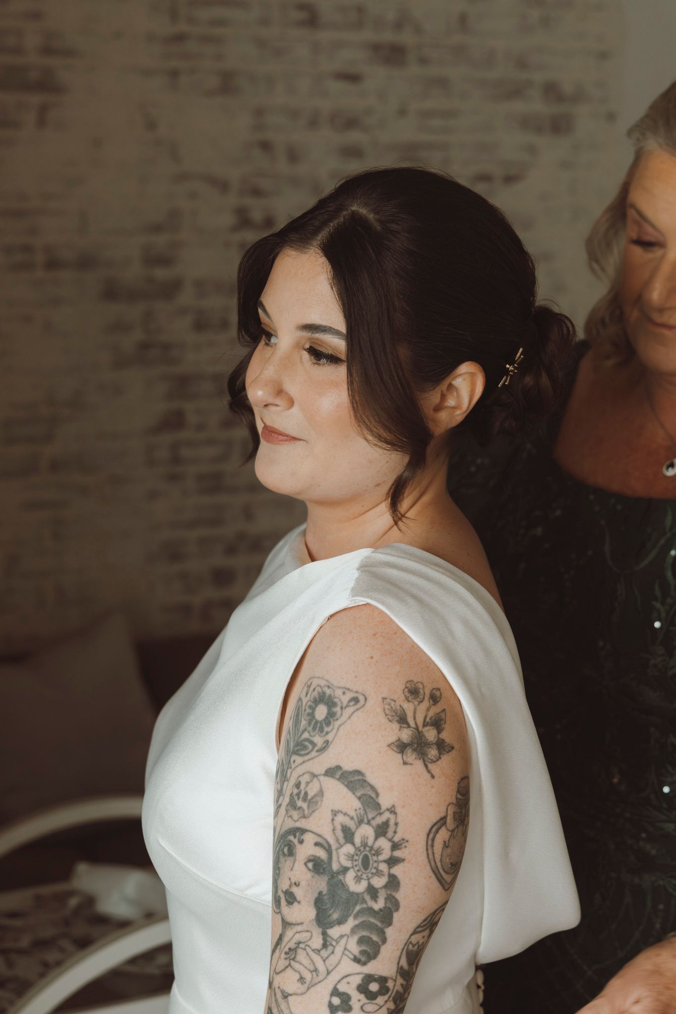 Close-up of person in white wedding dress with floral tattoo sleeve against brick wall.