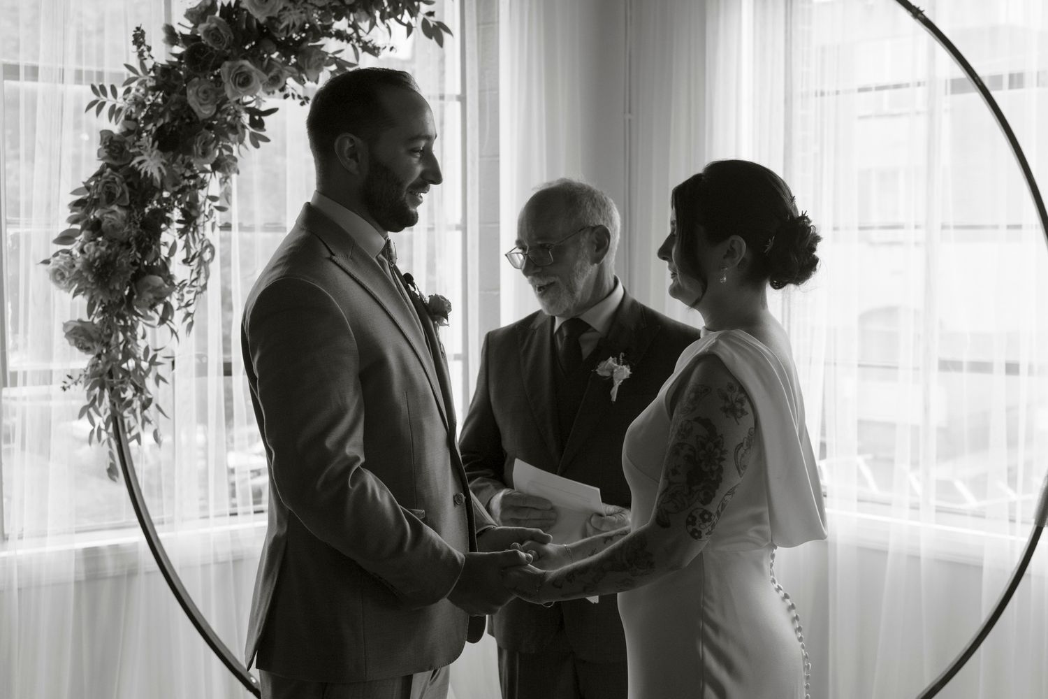 Black and white photo of a wedding ceremony with guests gathered in a bright room with large windows.