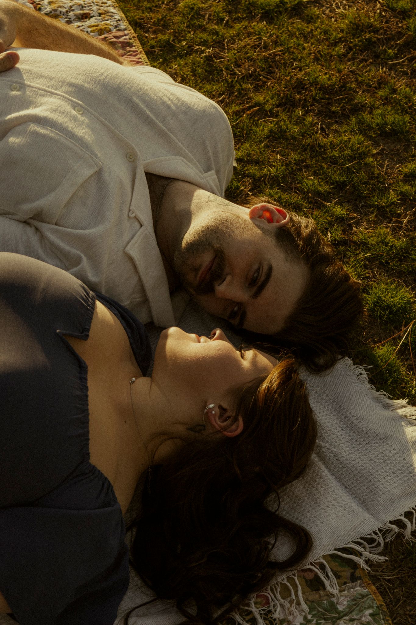 Couple lying on a blanket in sunlit grass, sharing an intimate moment face to face outdoors.
