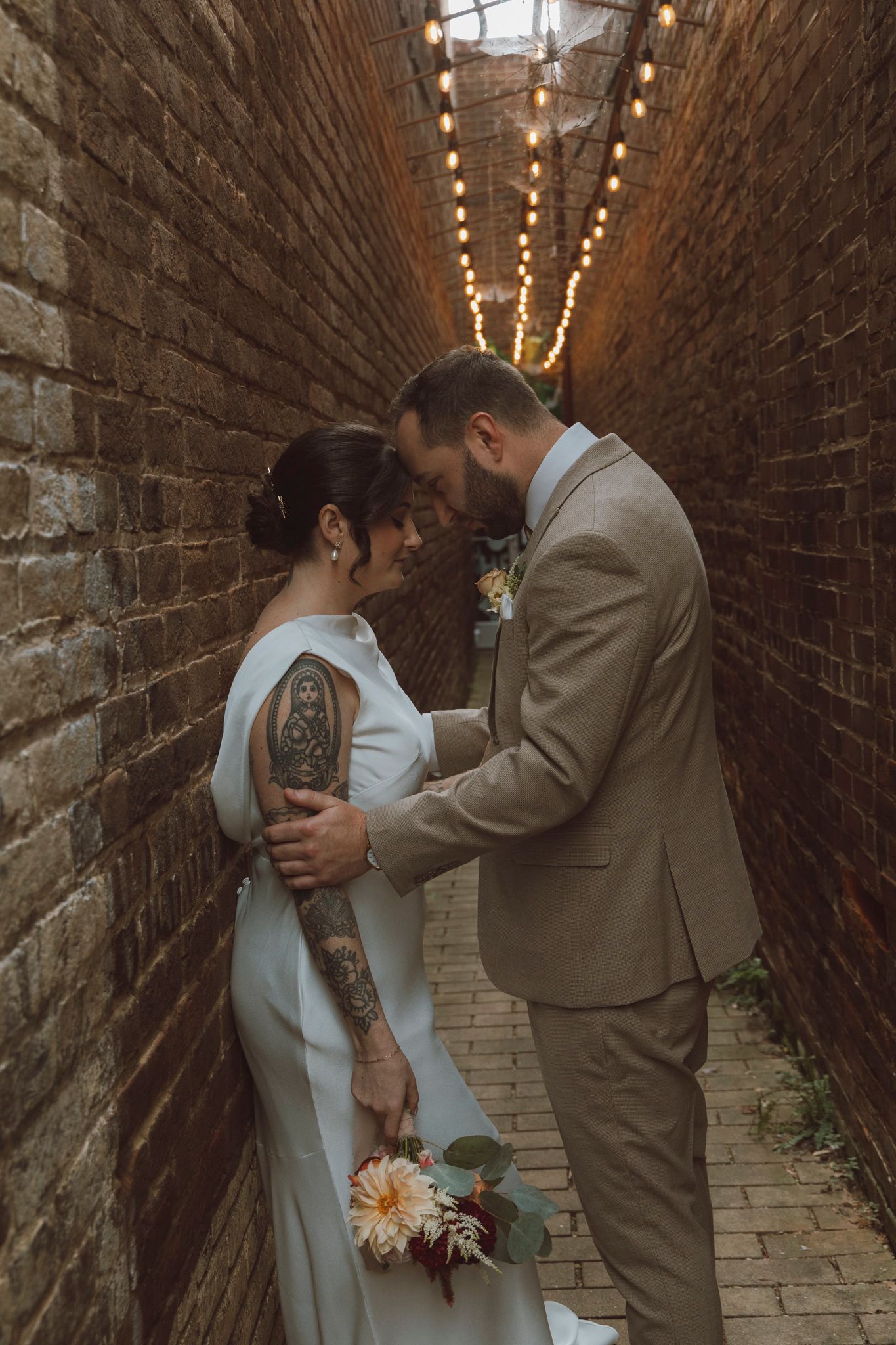 A romantic moment captured in a brick alley with string lights overhead as a couple shares an intimate wedding day embrace.