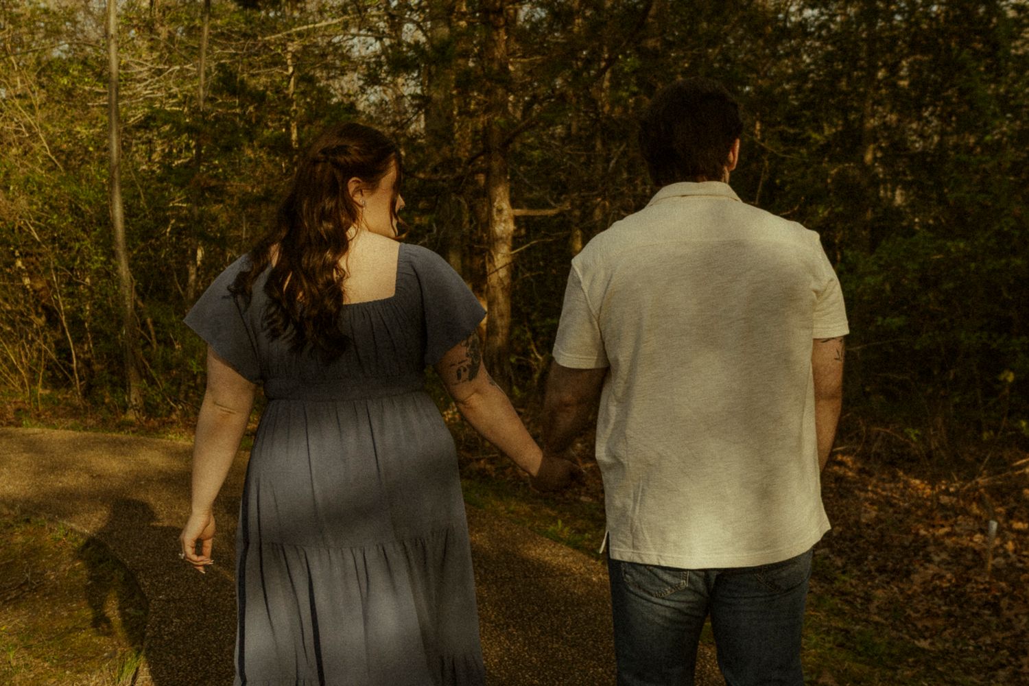 Couple walks hand in hand down a sunlit forest path, woman in blue dress and man in white shirt.