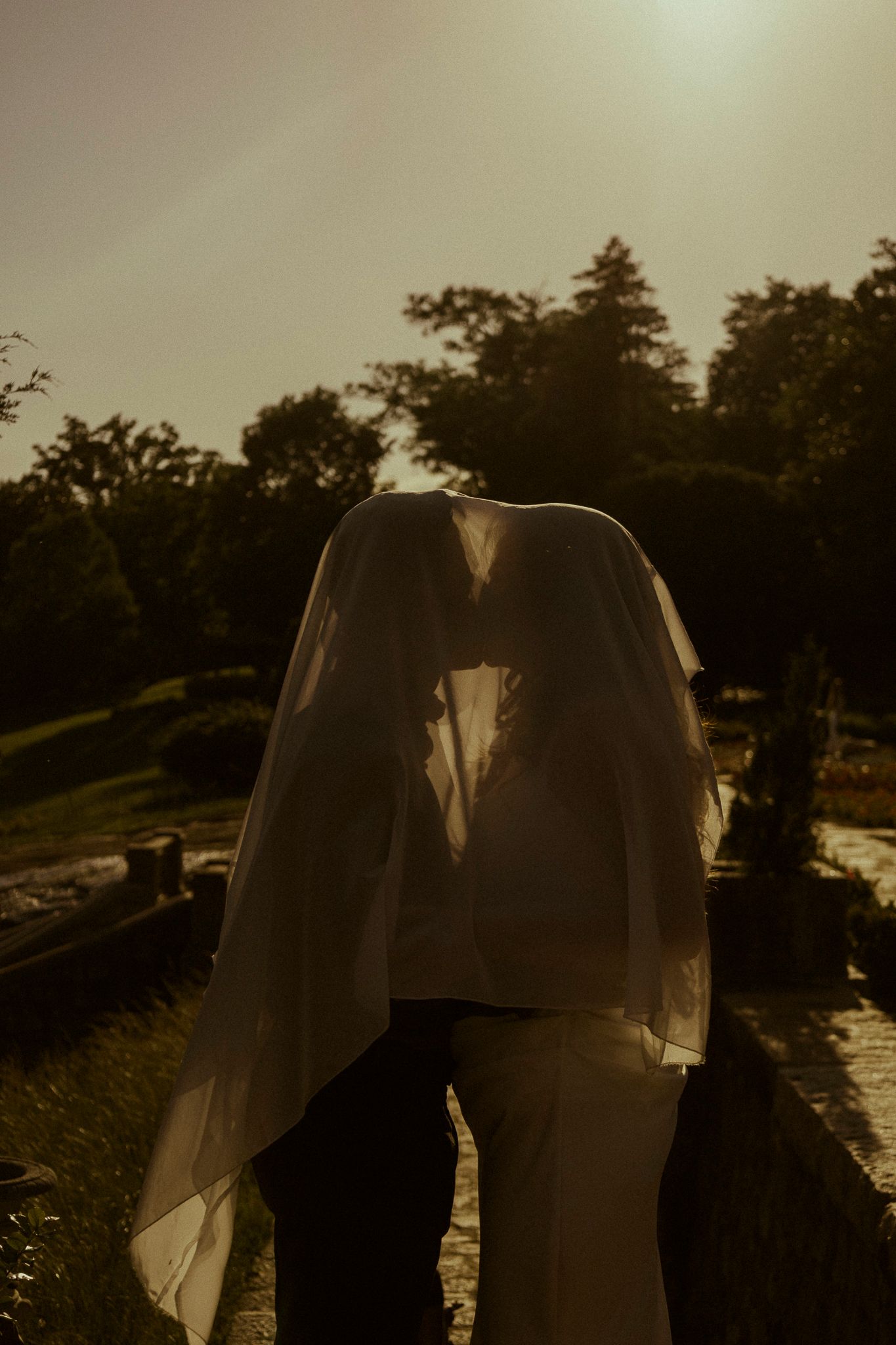 A bride and groom share a kiss under a wedding veil at golden hour in a lush garden setting.
