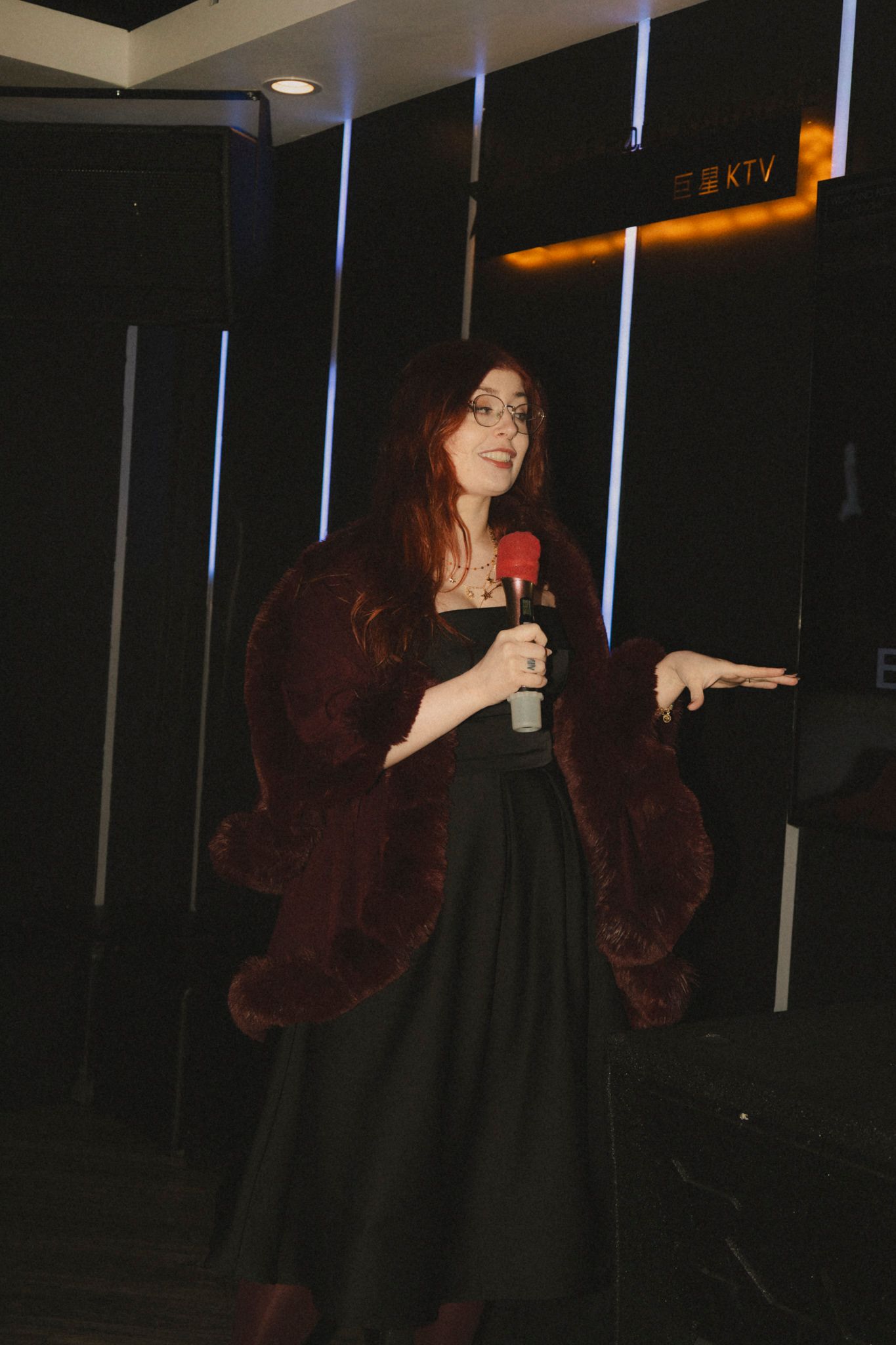 Woman in black dress and burgundy fur coat poses expressively in dark room with vertical light strips.
