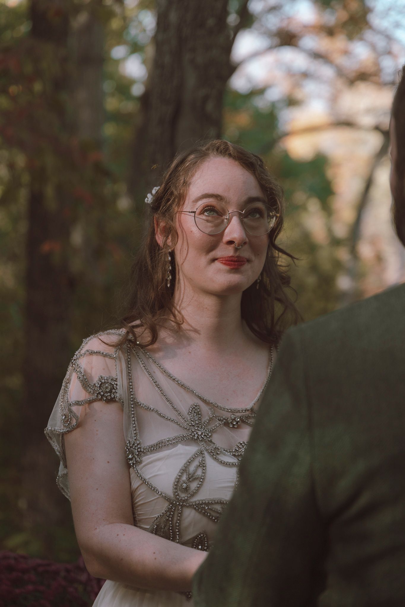 A person in vintage-inspired dress with glasses poses thoughtfully in a wooded outdoor setting during golden hour.