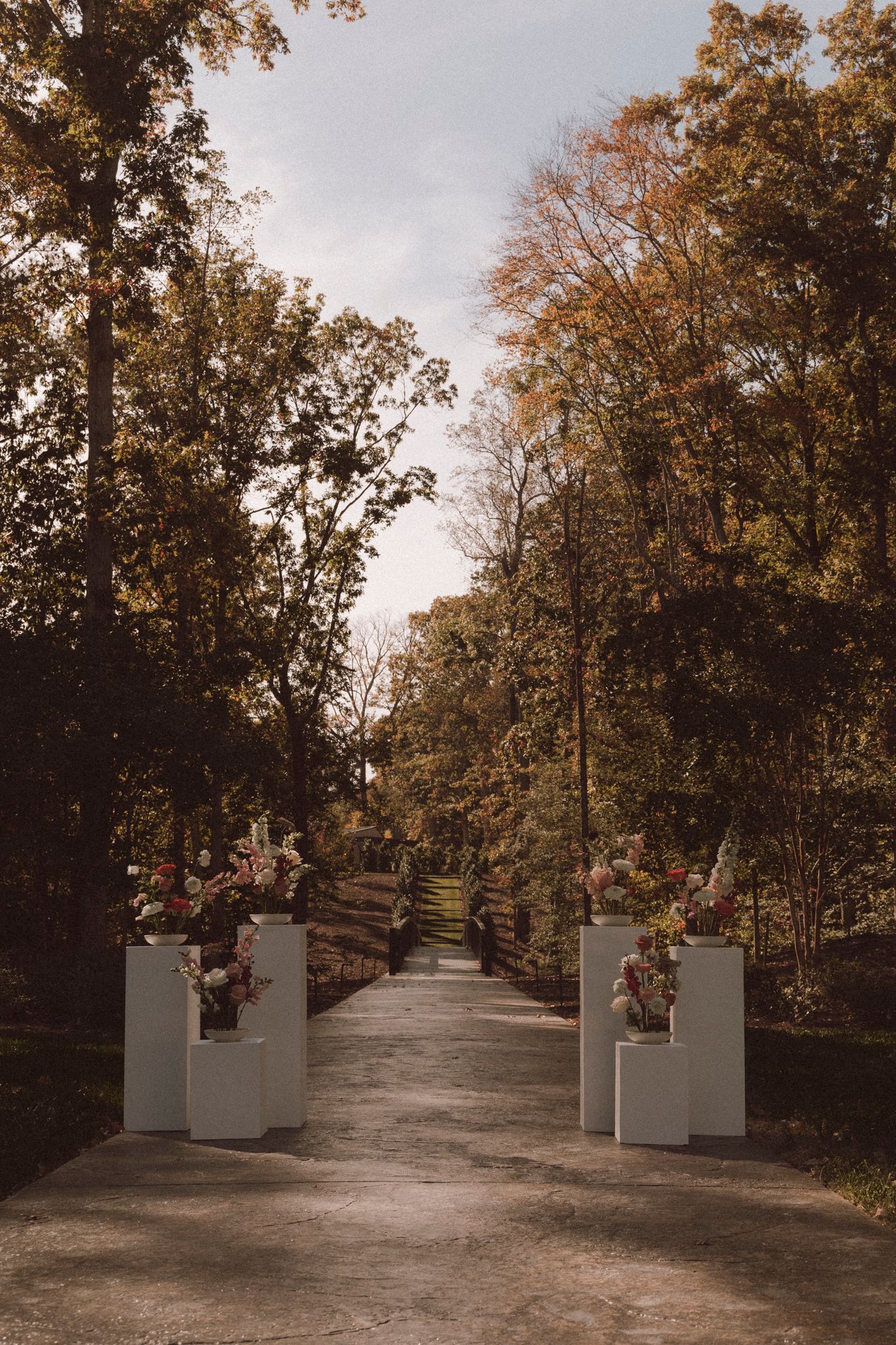 A curved garden path lined with white stone pillars and autumn trees with golden leaves creating a scenic walkway.