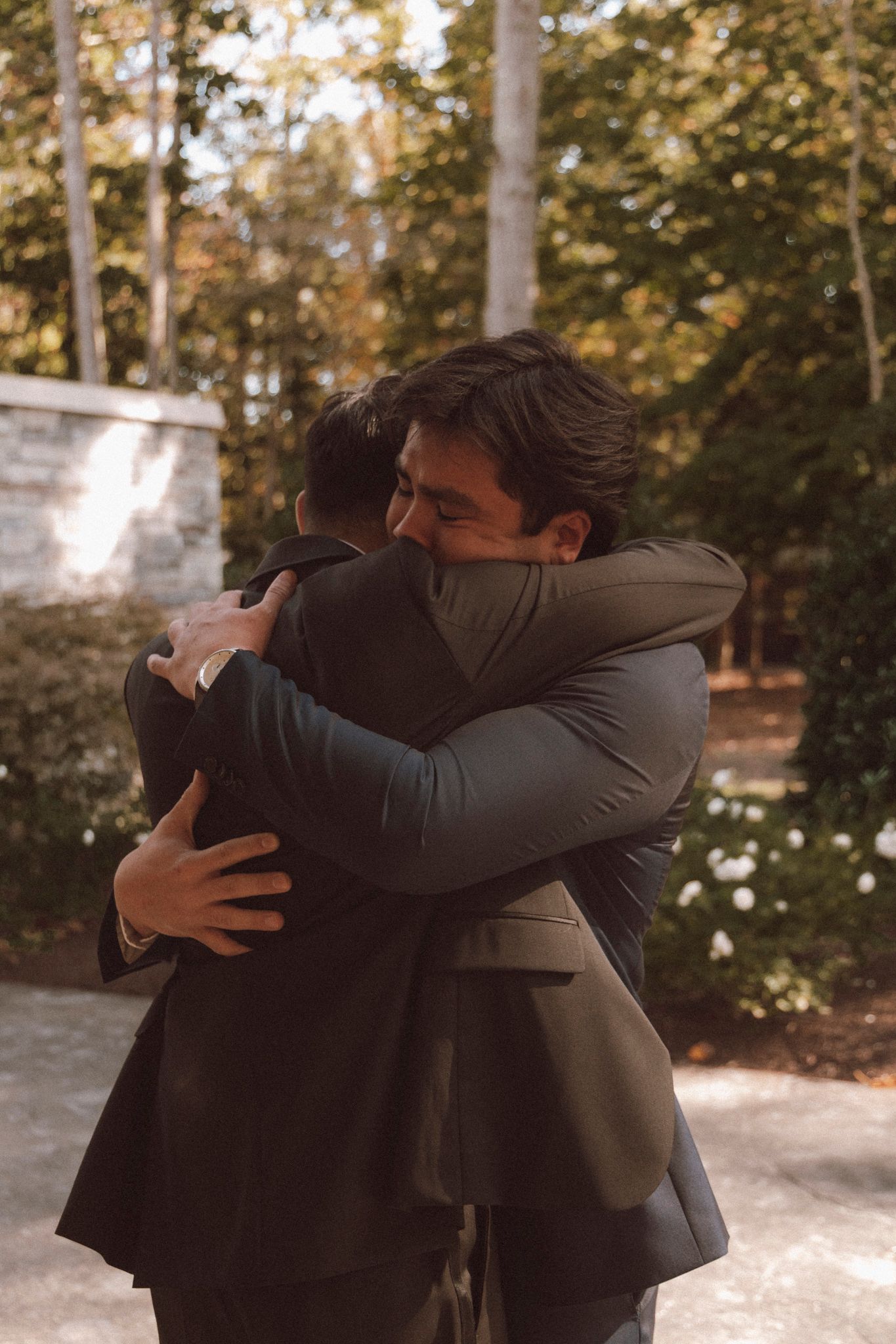 Two people in formal attire share an emotional embrace outdoors in a sunlit wooded setting.
