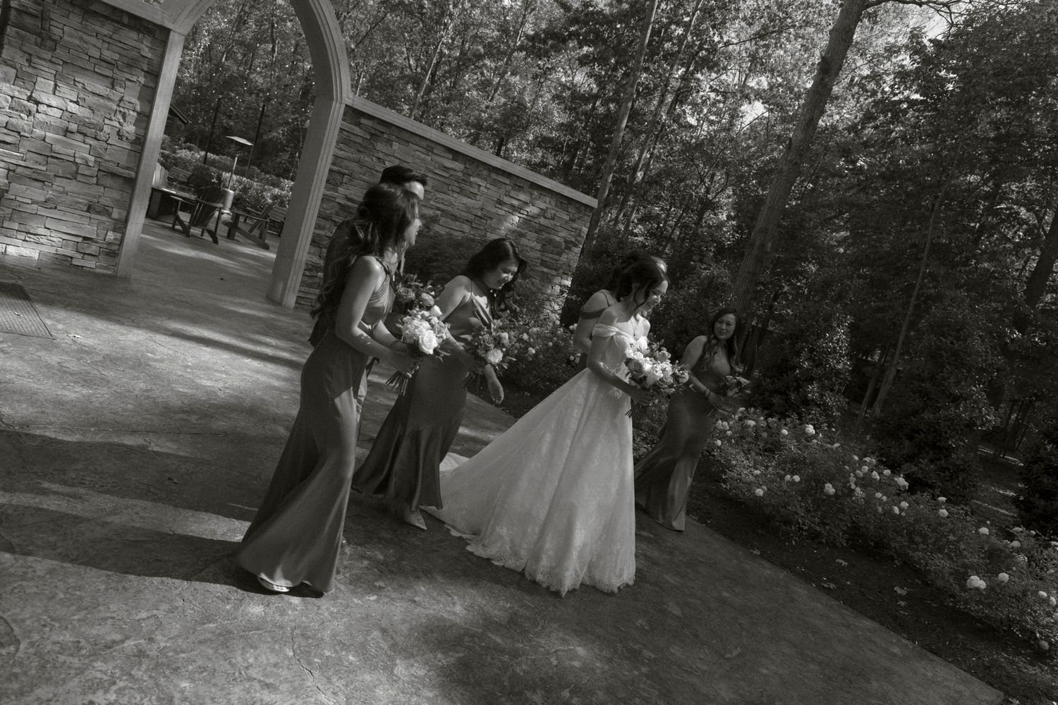 A black and white panoramic photo sequence shows a couple in formal attire walking through a wooded outdoor setting.