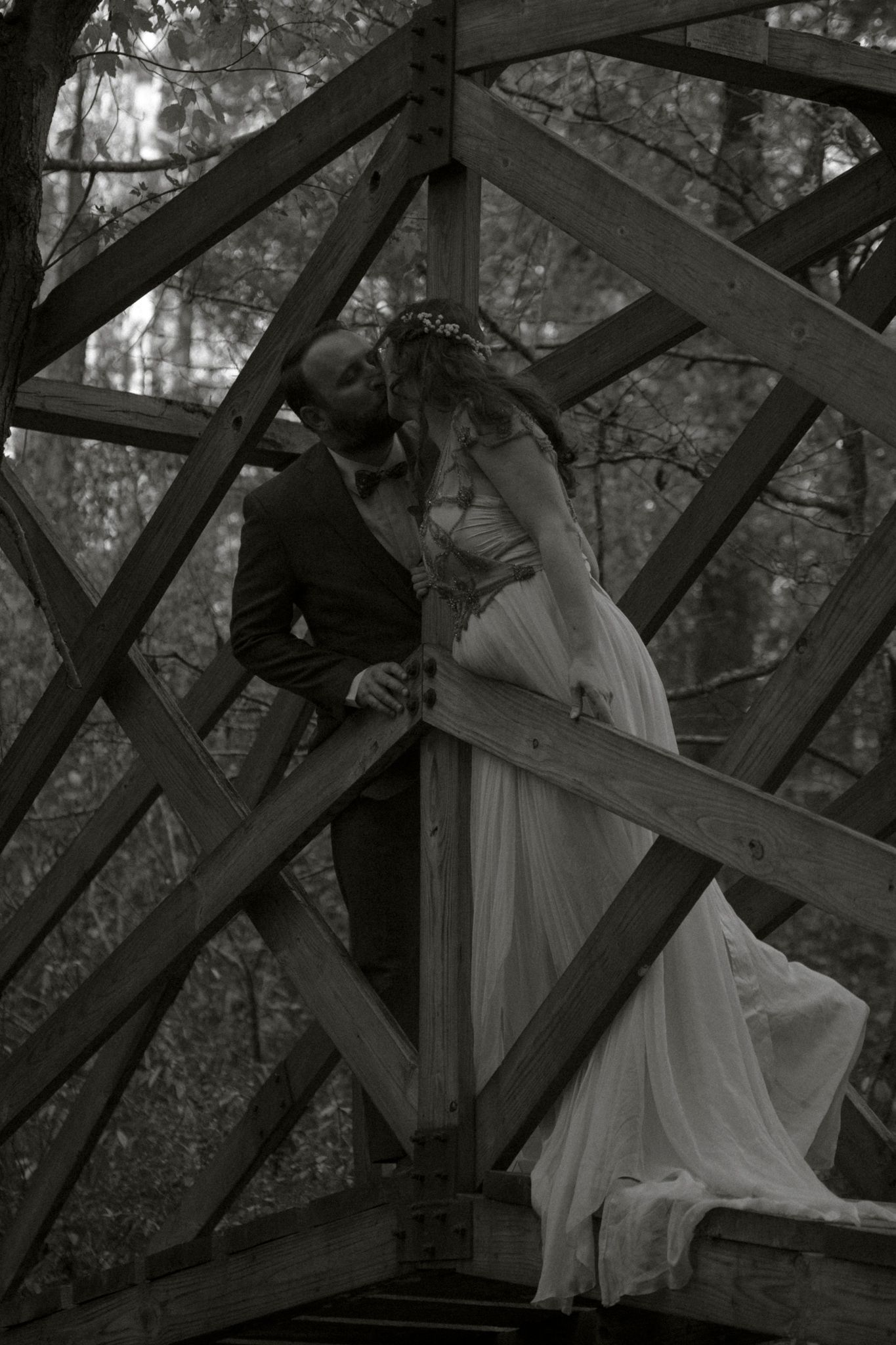 A figure in a flowing white dress poses elegantly against a rustic wooden bridge structure in a moody black and white photo.