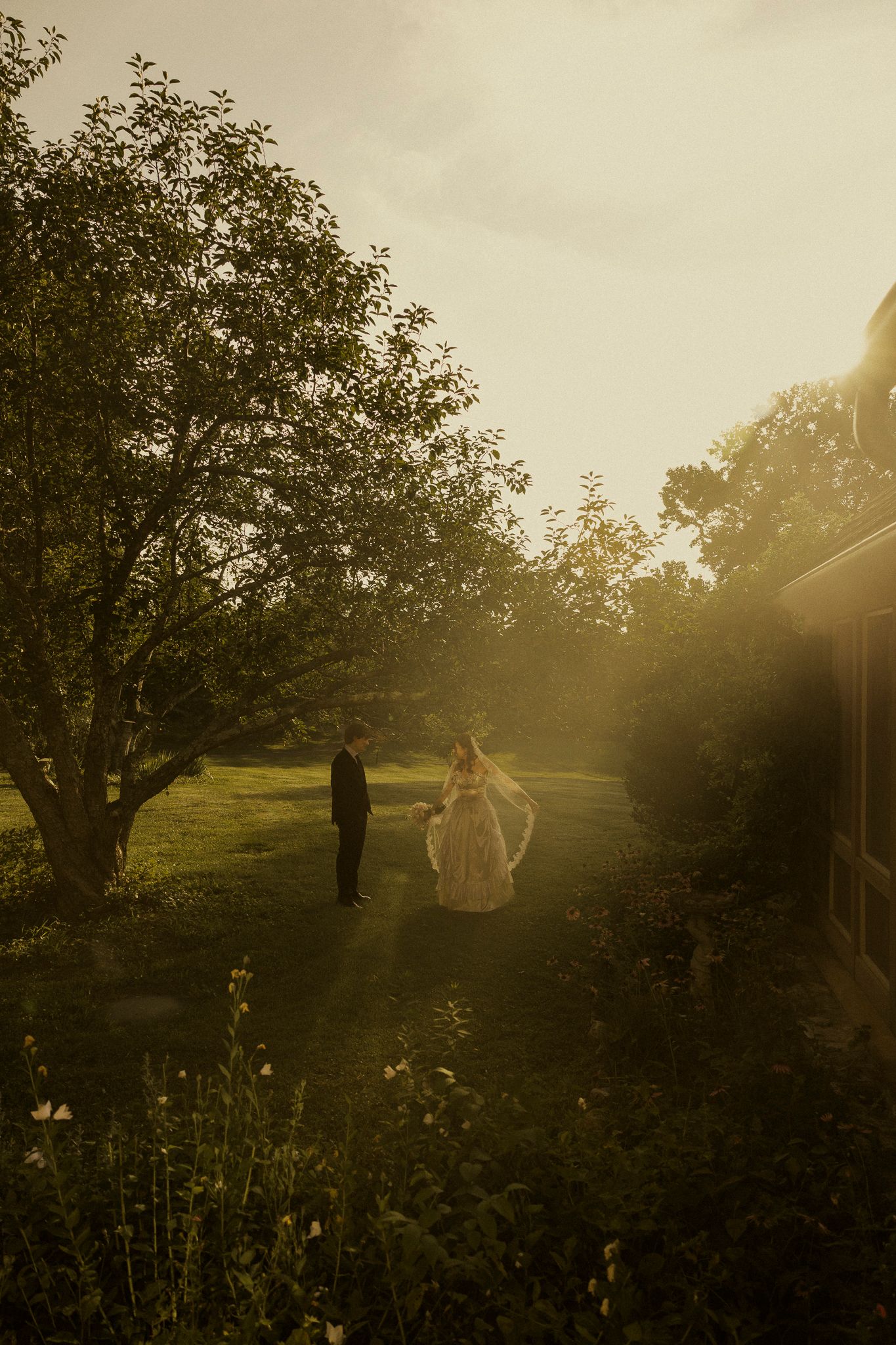 Bride and groom walking in golden sunset light among lush green trees at an outdoor wedding venue.