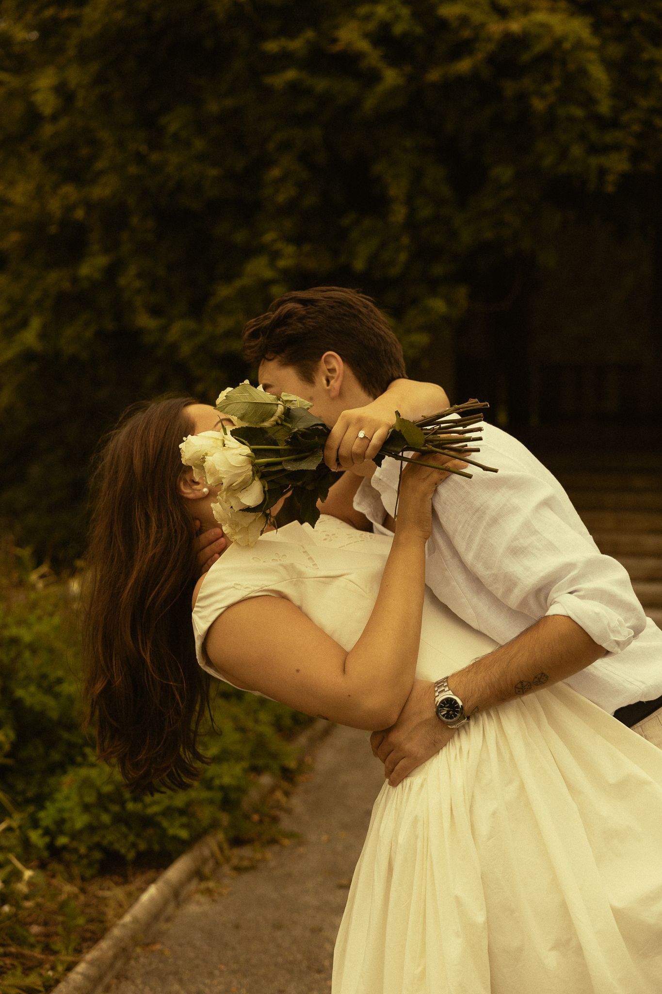 Romantic couple in white sharing a kiss while holding white flowers bouquet in a lush green garden setting.