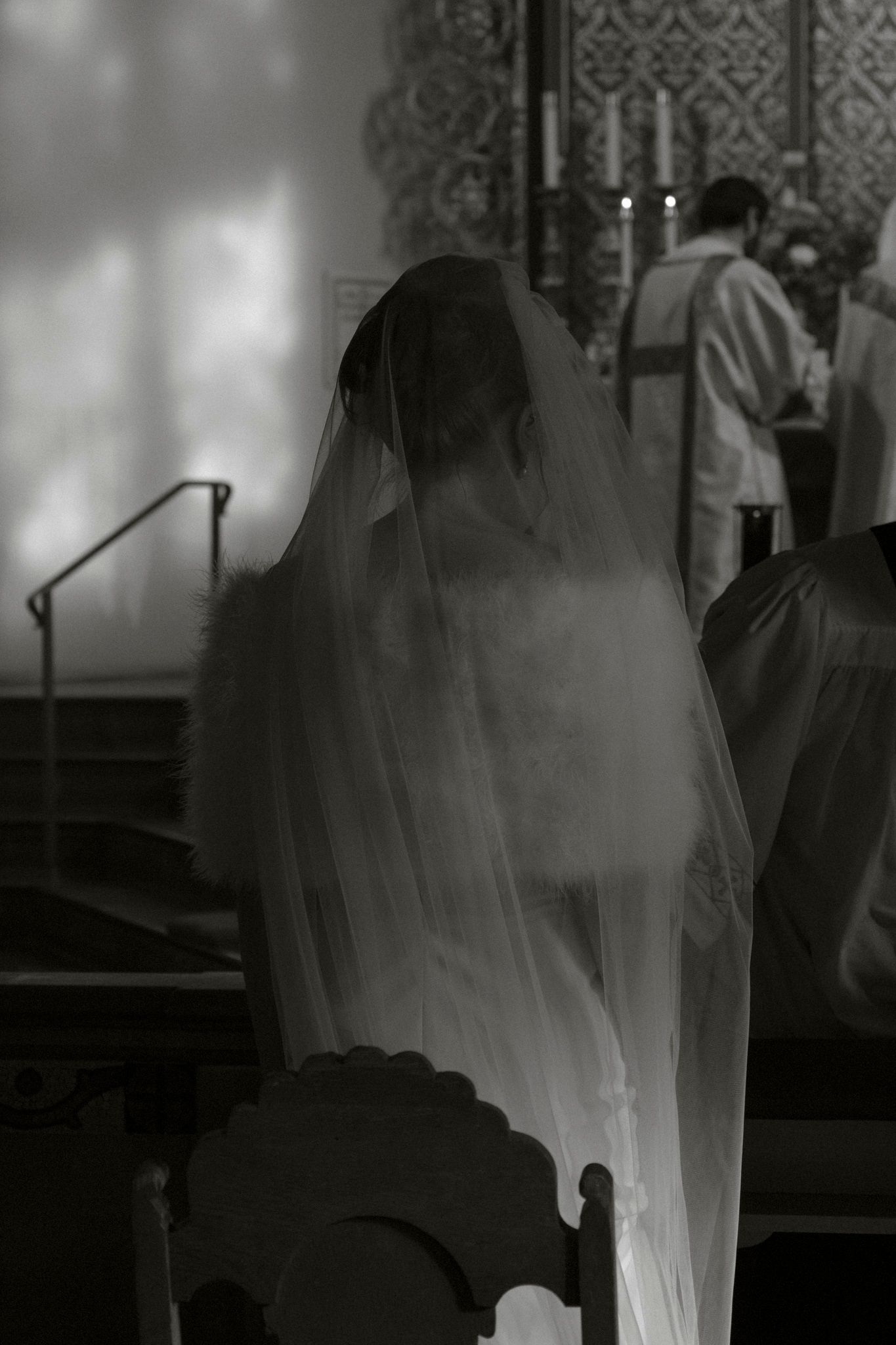 Black and white silhouette of bride in wedding veil seated at church altar during ceremony.