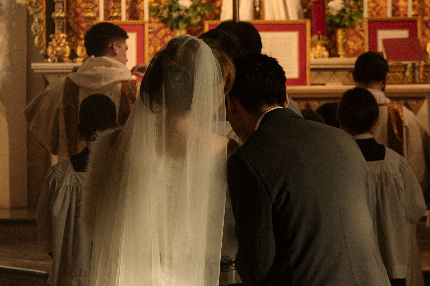 Bride in veil and groom kneel before priest at ornate church altar.