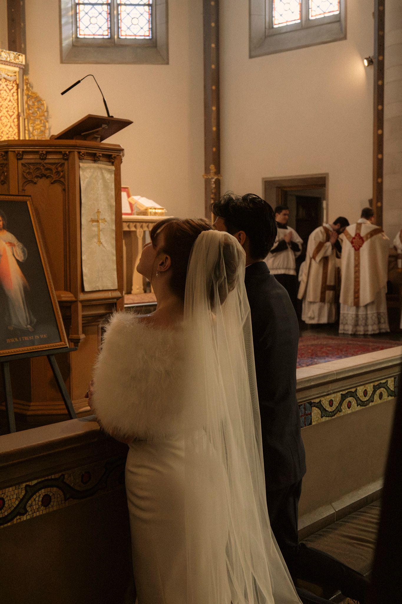 Bride in white fur-trimmed gown and cathedral veil kneeling beside groom before ornate Catholic church altar during ceremony.