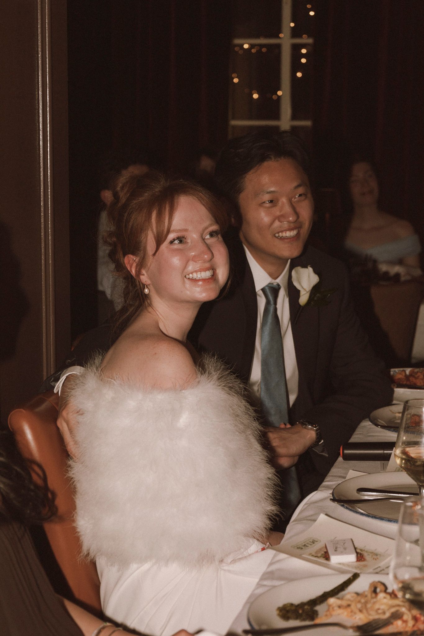Bride in white fur wrap and groom in dark suit smiling together at their wedding reception dinner table.