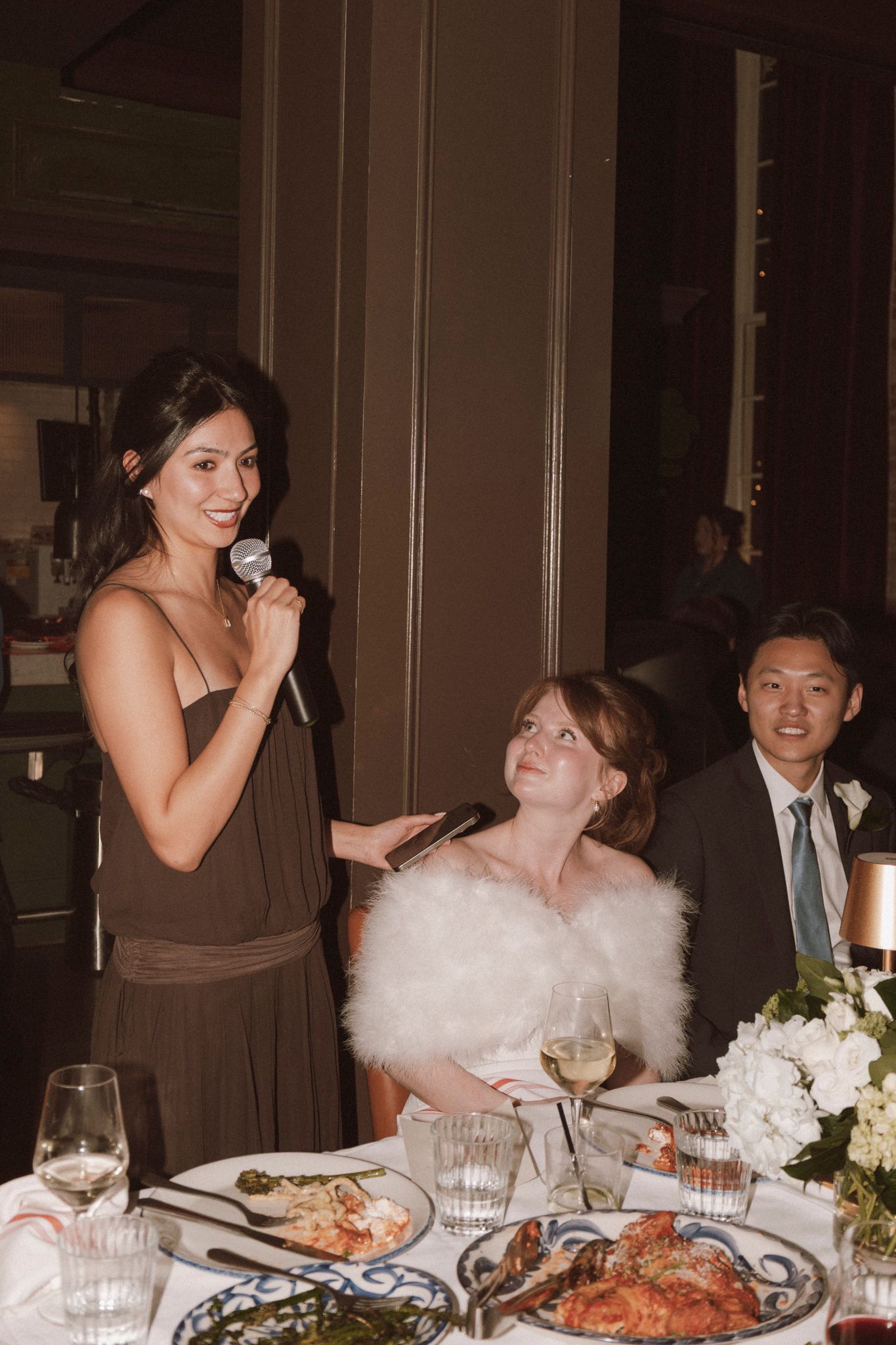 Bridesmaid in brown dress gives a toast with microphone while bride in white fur wrap listens smiling.