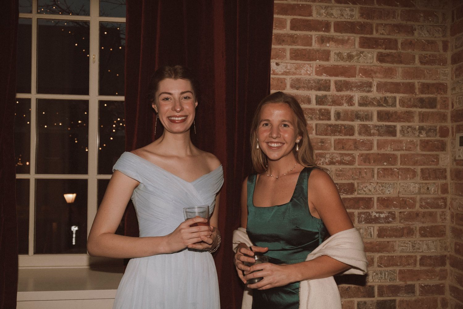 Two elegant women smiling at a formal event, one in a white gown and one in a green dress, holding drinks.
