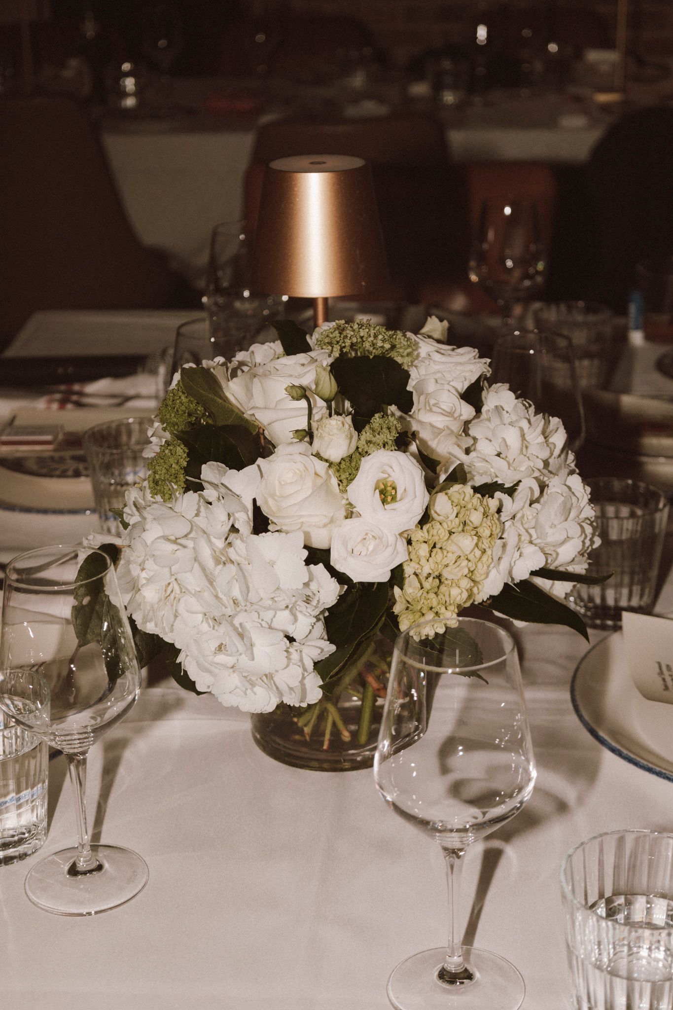 Elegant white floral centerpiece with roses and hydrangeas on a white tablecloth at a formal dinner.