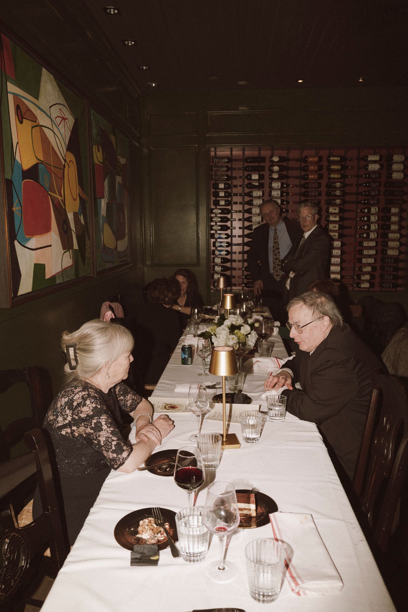 Elegantly dressed guests dine at long white-clothed table in upscale restaurant with wine wall and modern art.