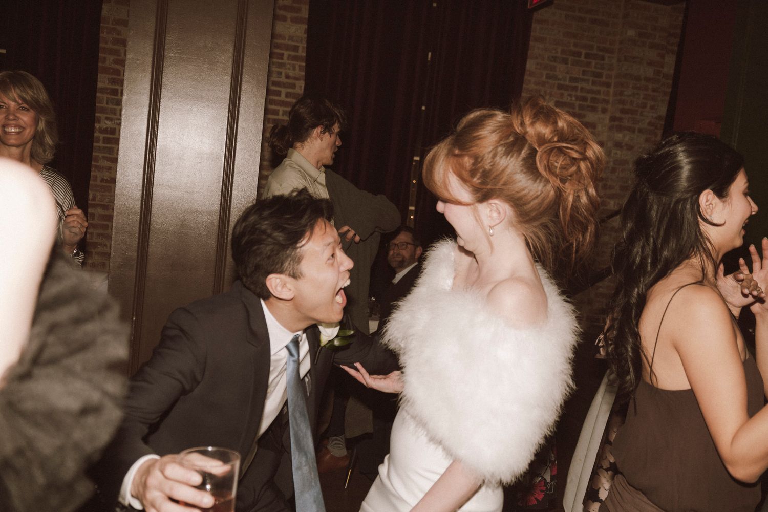 Bride in white fur stole dances with groom at lively wedding reception in a warmly lit venue.