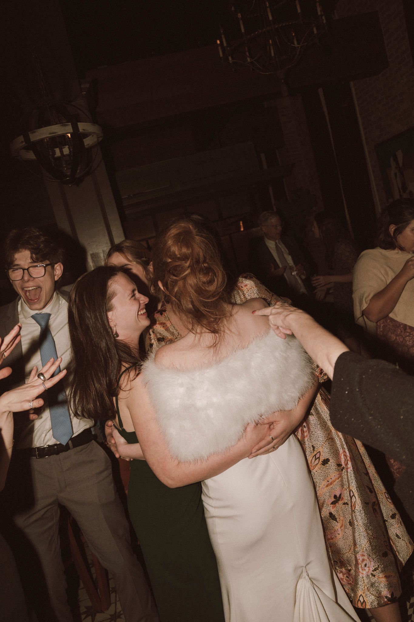 Bride in white gown with fur stole embracing a guest on the wedding reception dance floor.