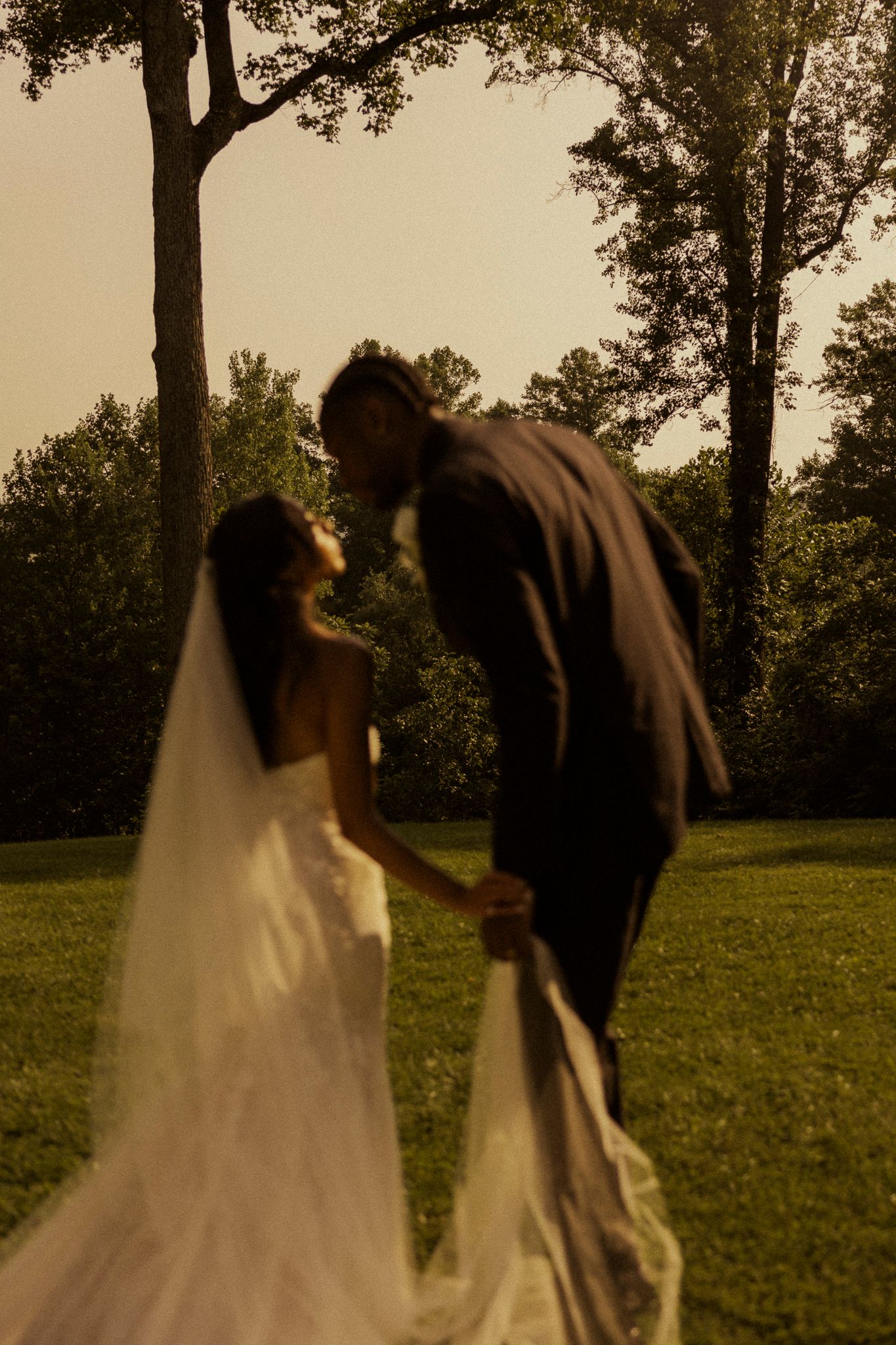 A bride and groom share a romantic moment outdoors, holding hands in a lush green garden at golden hour.