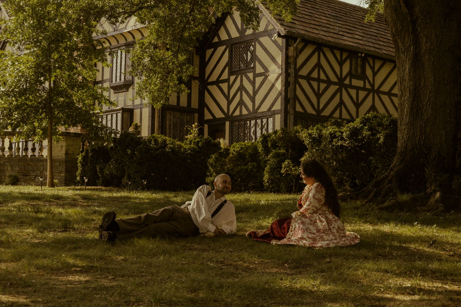 Couple in vintage attire relaxing on grass beneath a tree near a Tudor-style building.