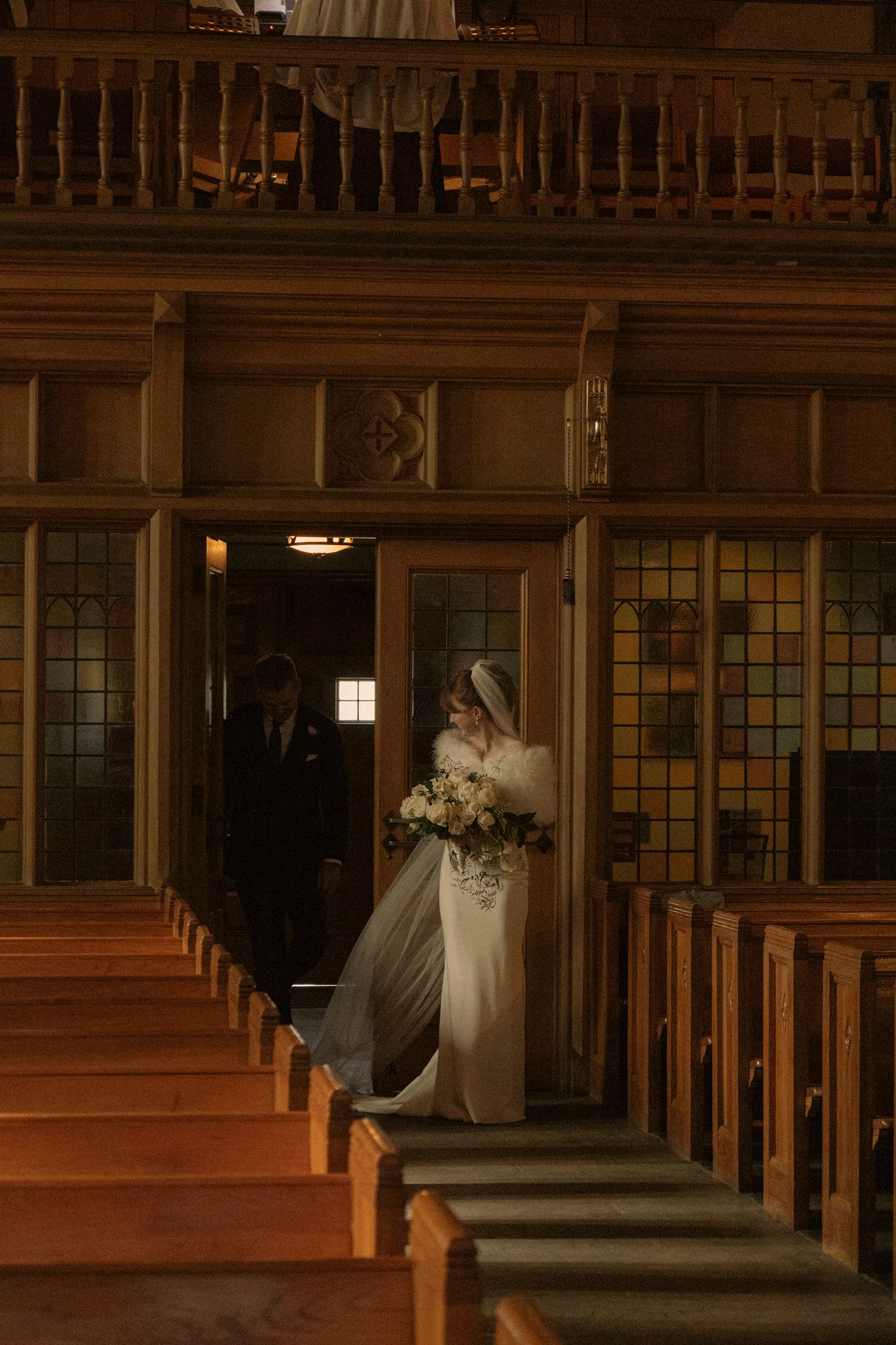 Bride in white gown and veil holds bouquet at church entrance with groom behind her, wooden pews lining the aisle.