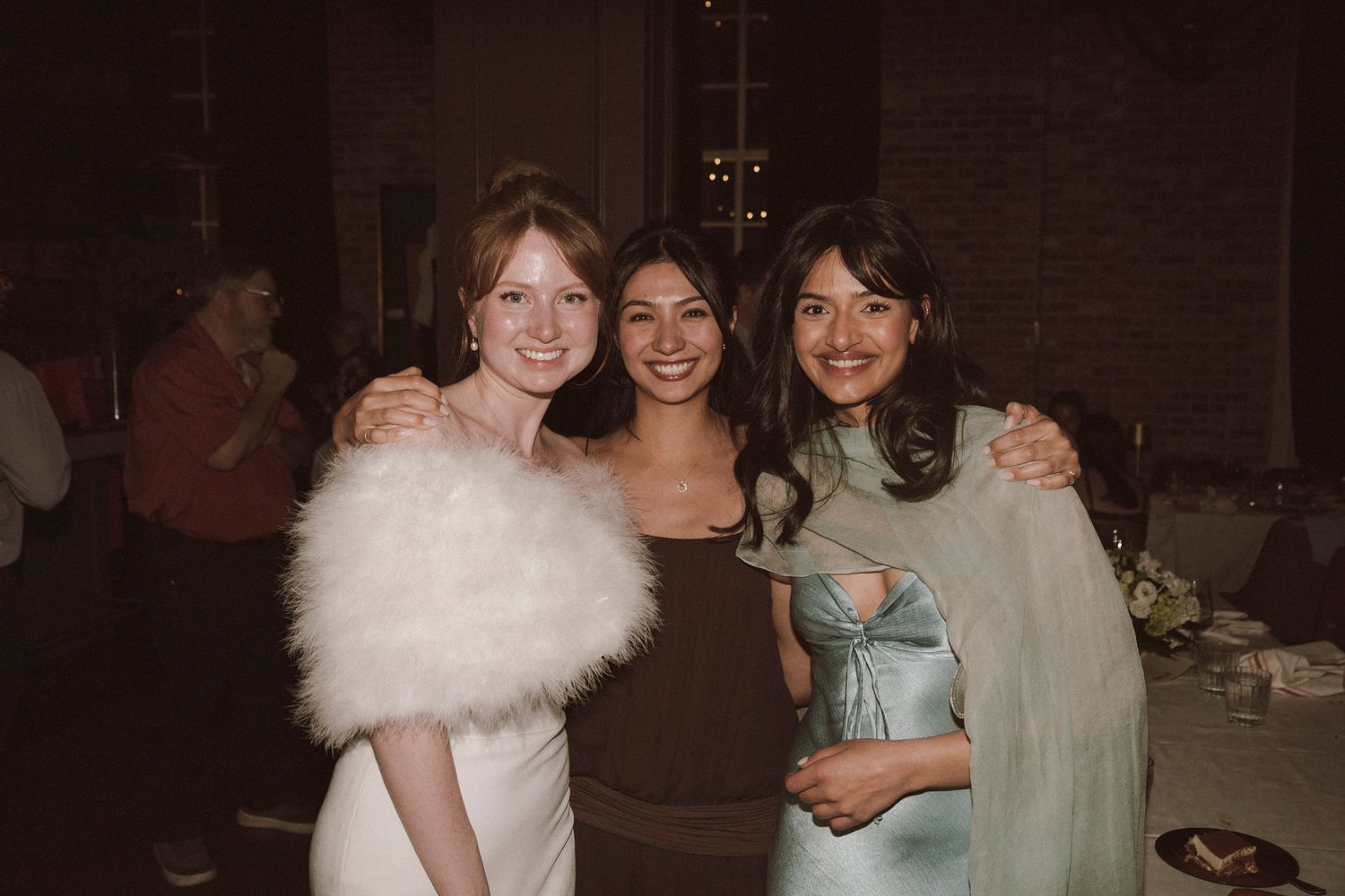 Three women in formal attire posing together at a wedding reception venue.