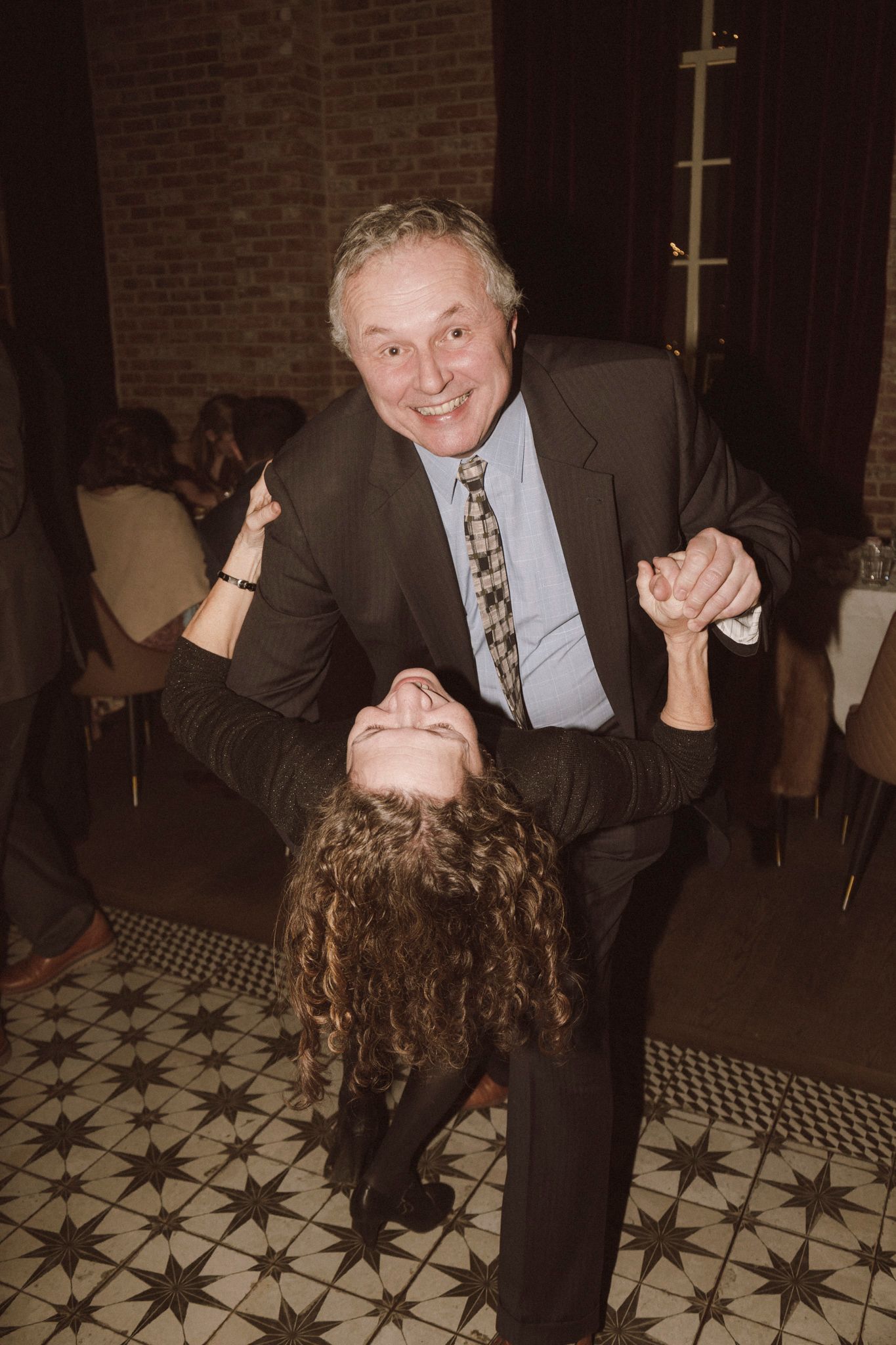 An older man in a suit dips a curly-haired woman on a patterned dance floor at a formal event.
