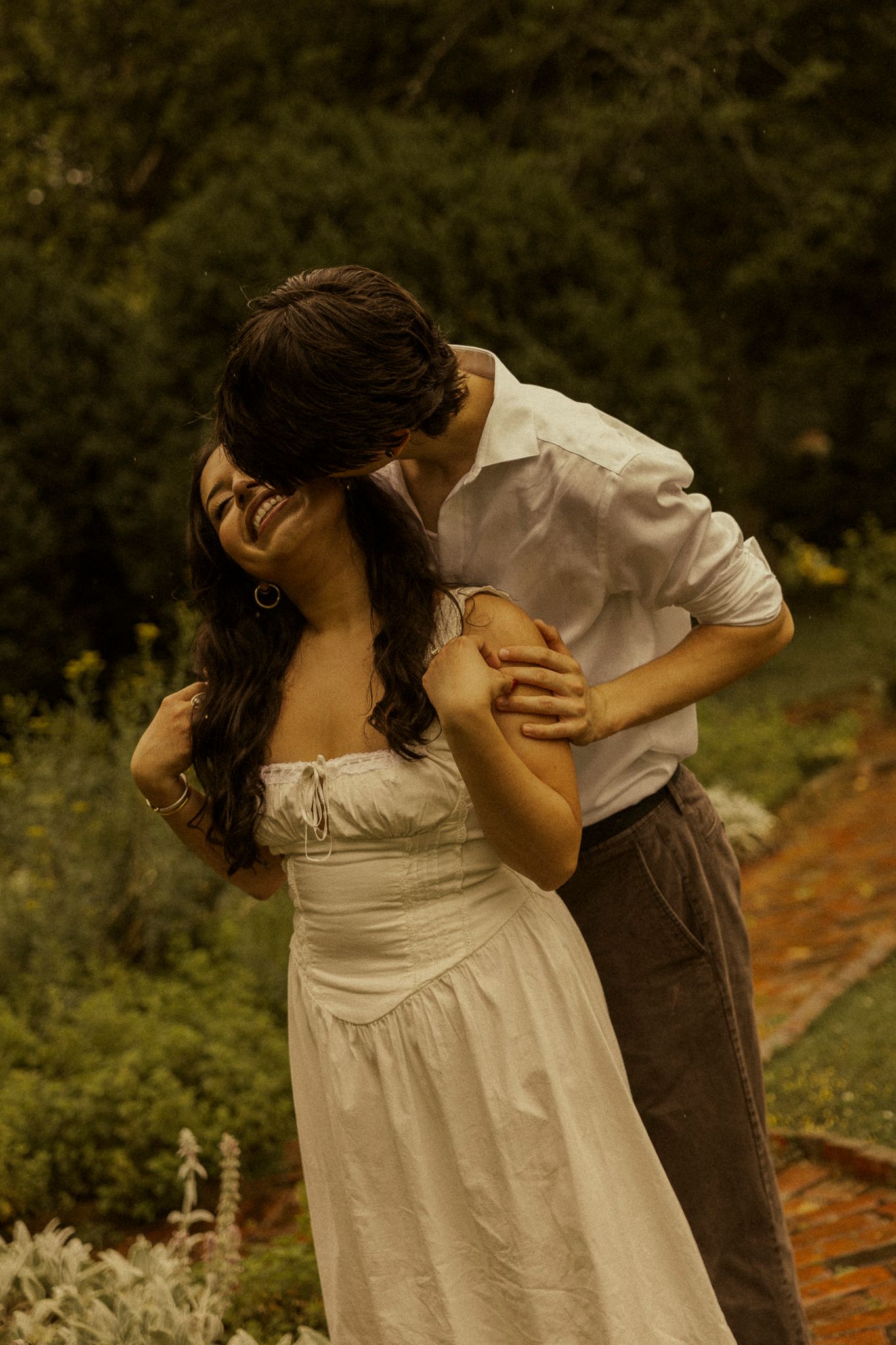 A couple shares a romantic embrace in a lush garden, the woman in a white dress laughing joyfully.