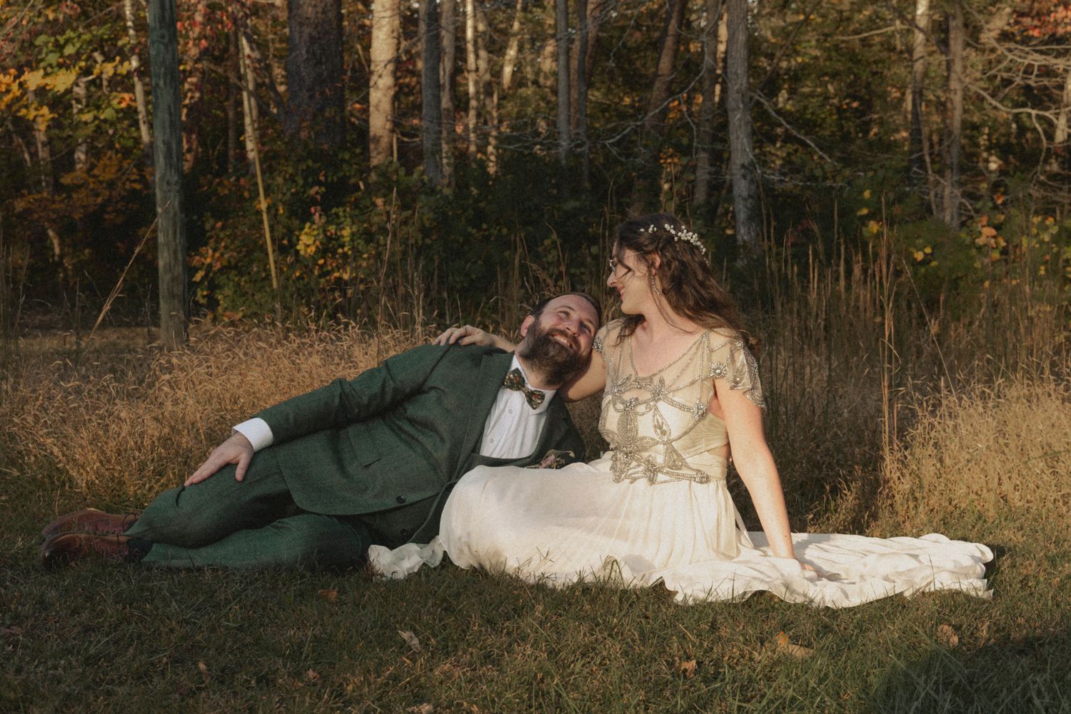 A couple in wedding attire lies together on autumn grass surrounded by golden trees at sunset.