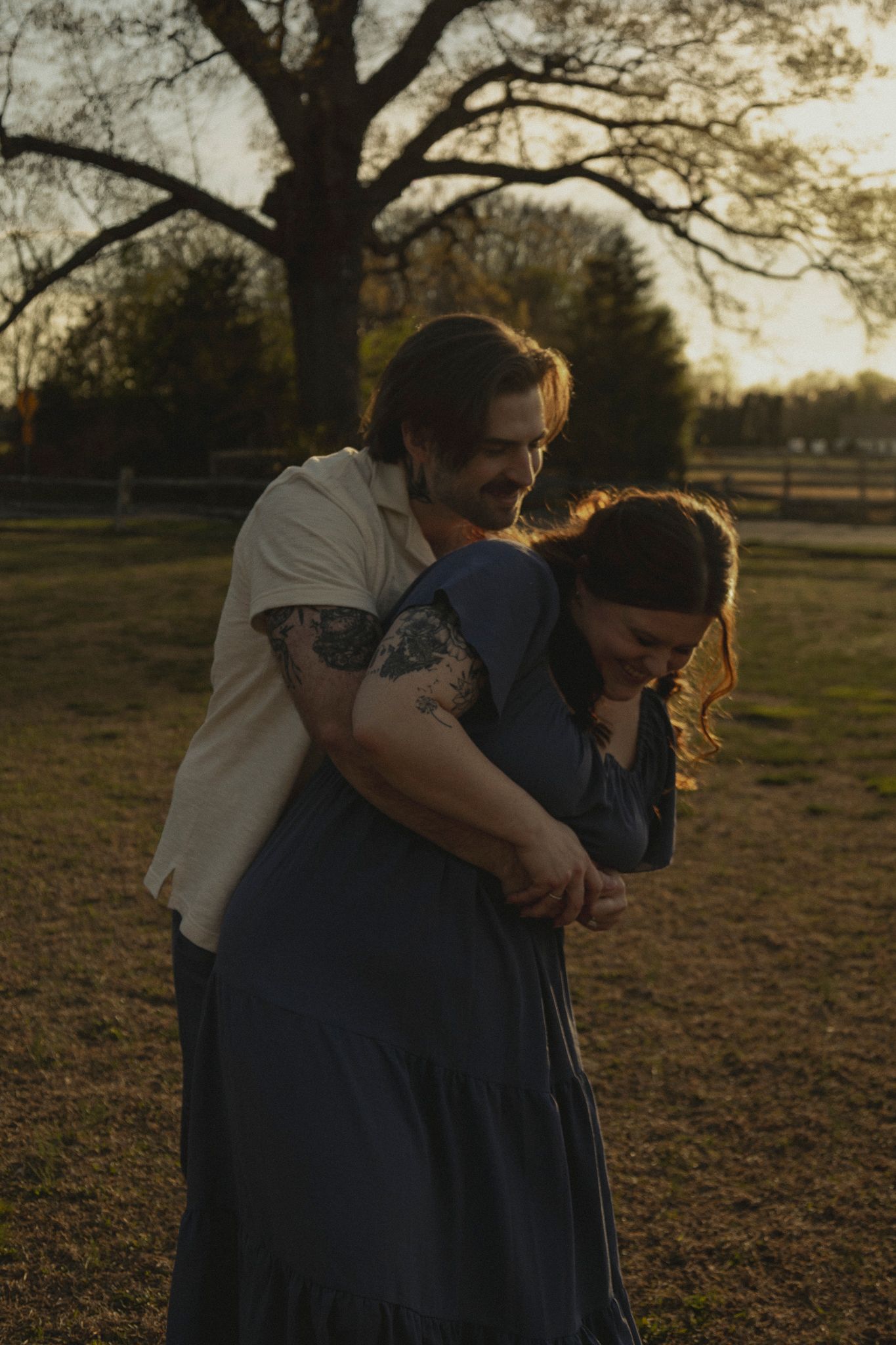 Couple laughing and embracing at golden hour in a park, man with tattoos hugging woman in blue dress.